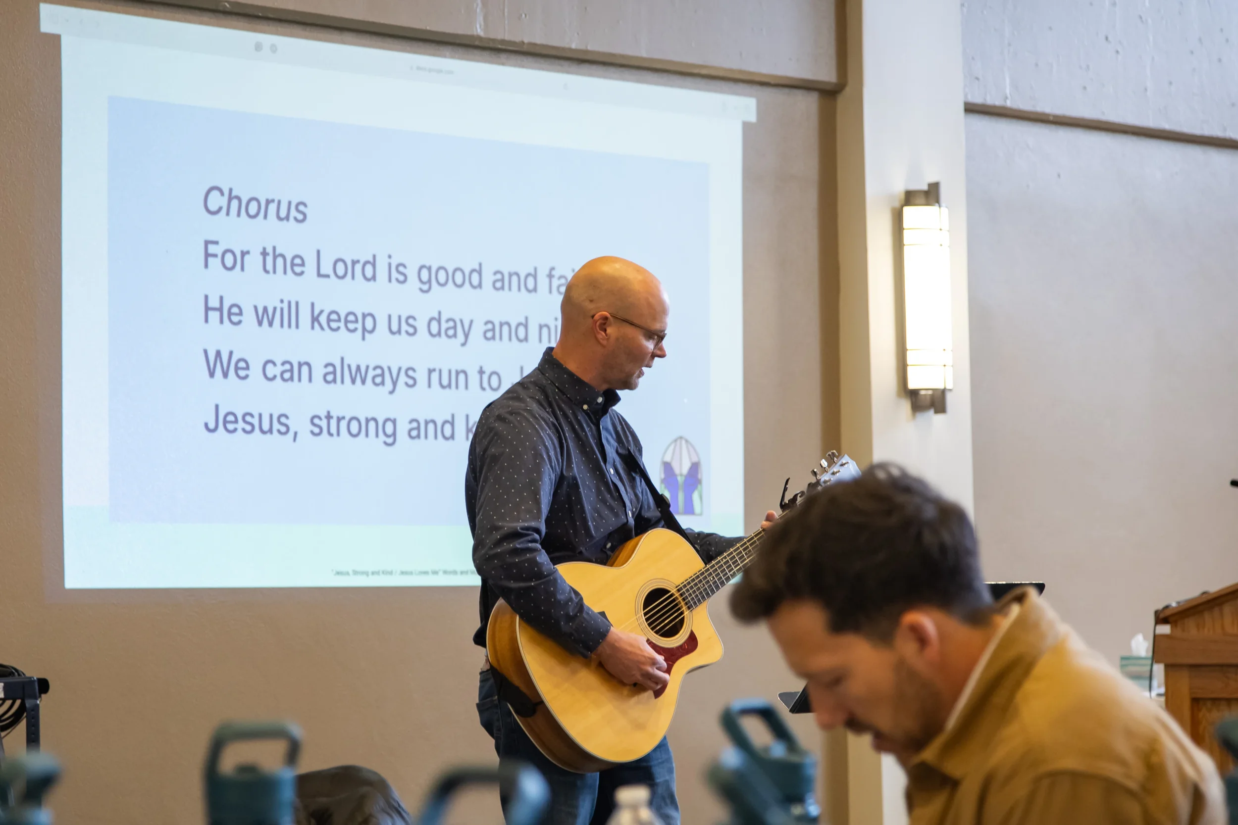 Worship leader playing acoustic guitar during Teach Us to Worship conference with hymn lyrics projected on screen.