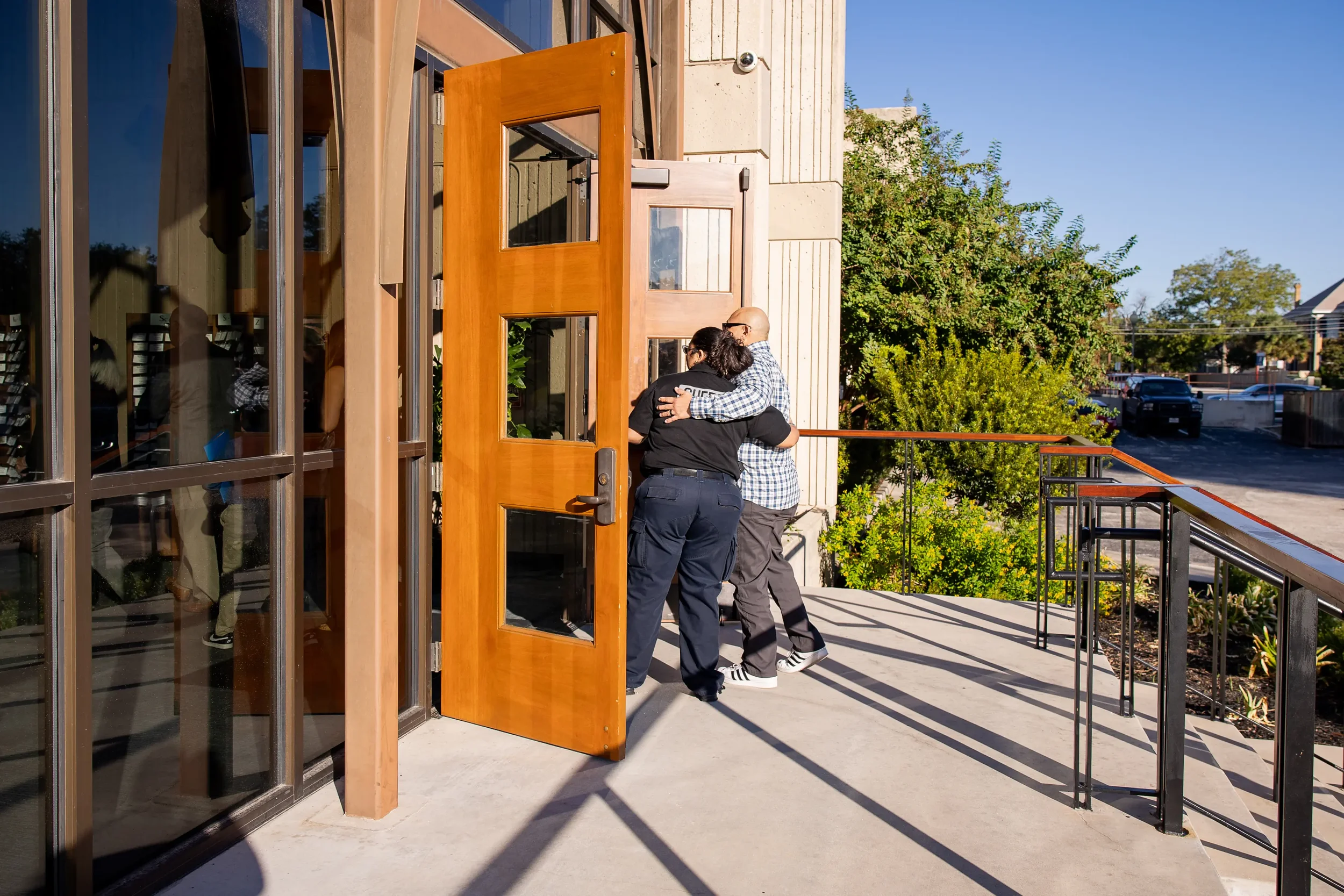 Two church attendees embracing while entering a church building through open wooden doors on a sunny day.