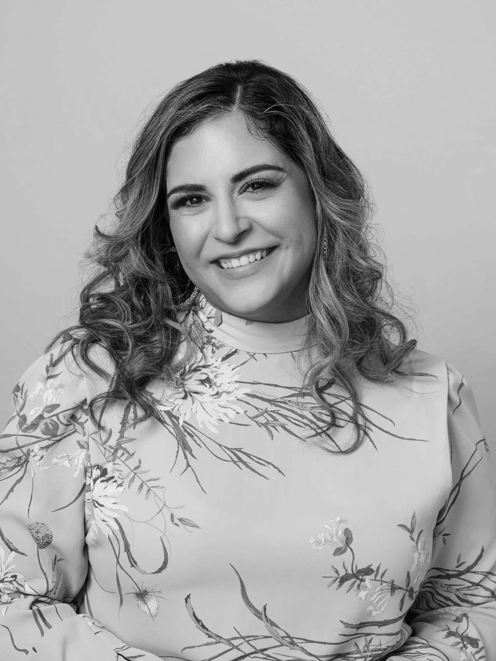 Professional headshot of a woman in a floral blouse and light jeans, smiling against a neutral background.