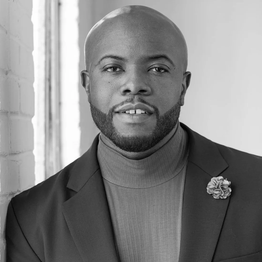 Black and white professional portrait of a man in a suit jacket with a flower lapel pin, leaning against a window with hands folded.
