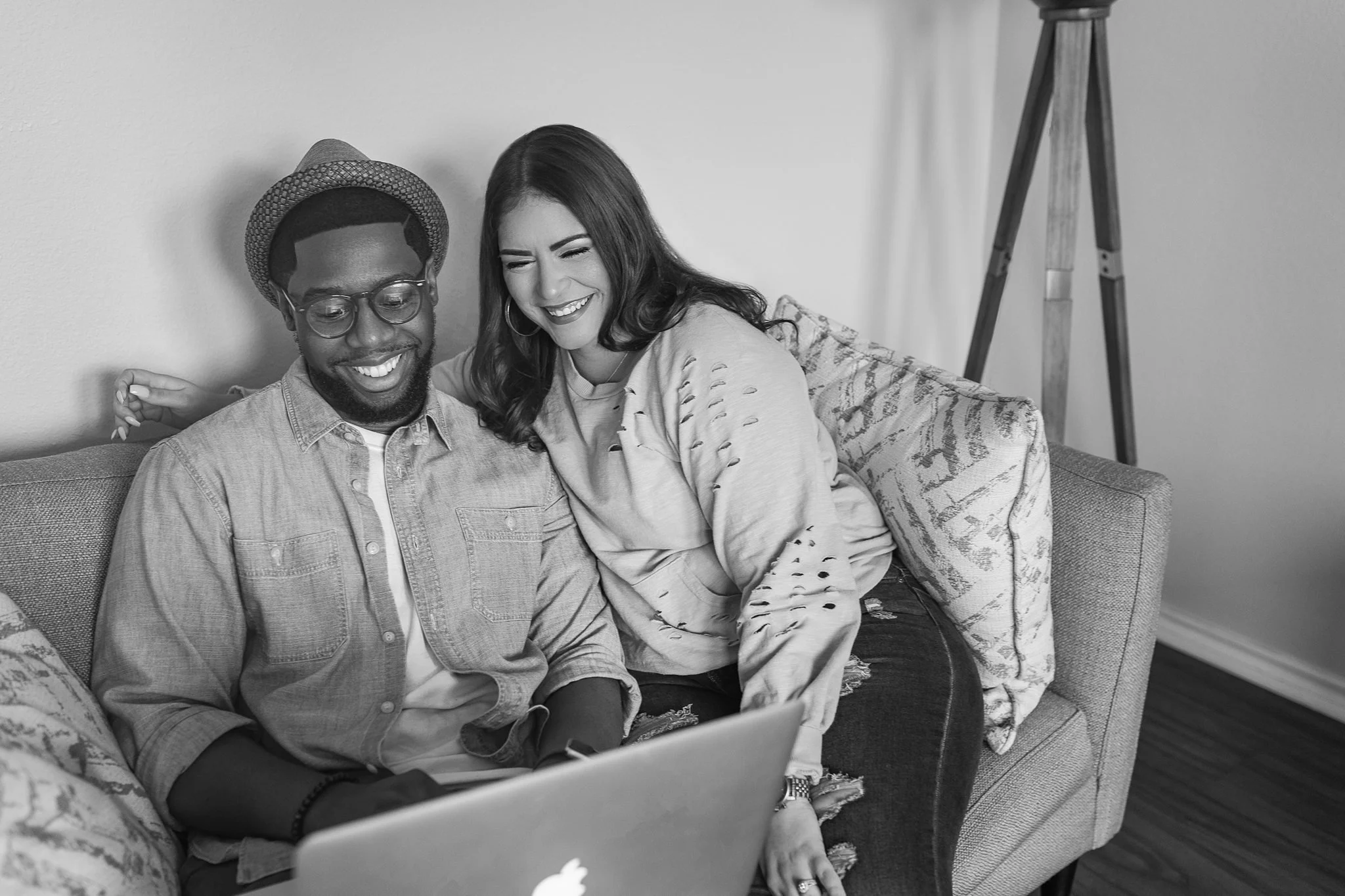 Black and white lifestyle photo of Jason and Alejandra Bryant smiling on a couch while working together on a laptop at home.