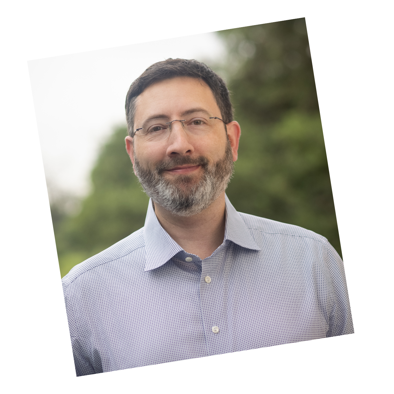Business headshot of a man with beard and glasses wearing a checkered shirt, outdoors with trees in the background.