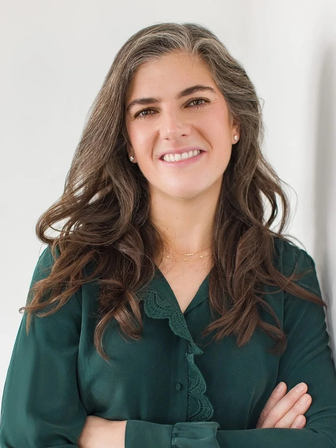 A woman with long brown wavy hair smiling with arms crossed, wearing a dark green blouse, earrings, and a delicate necklace, standing in front of a light background.