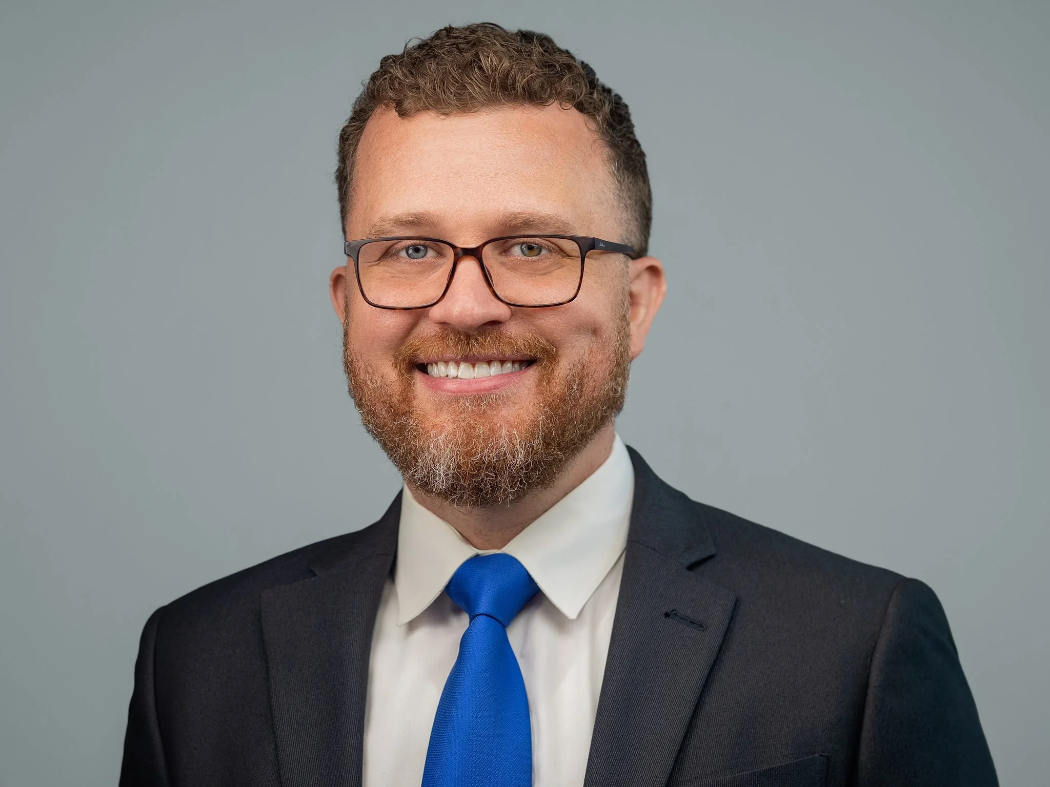 Business professional headshot of a man in a suit with a bright blue tie and glasses, smiling against a gray backdrop.