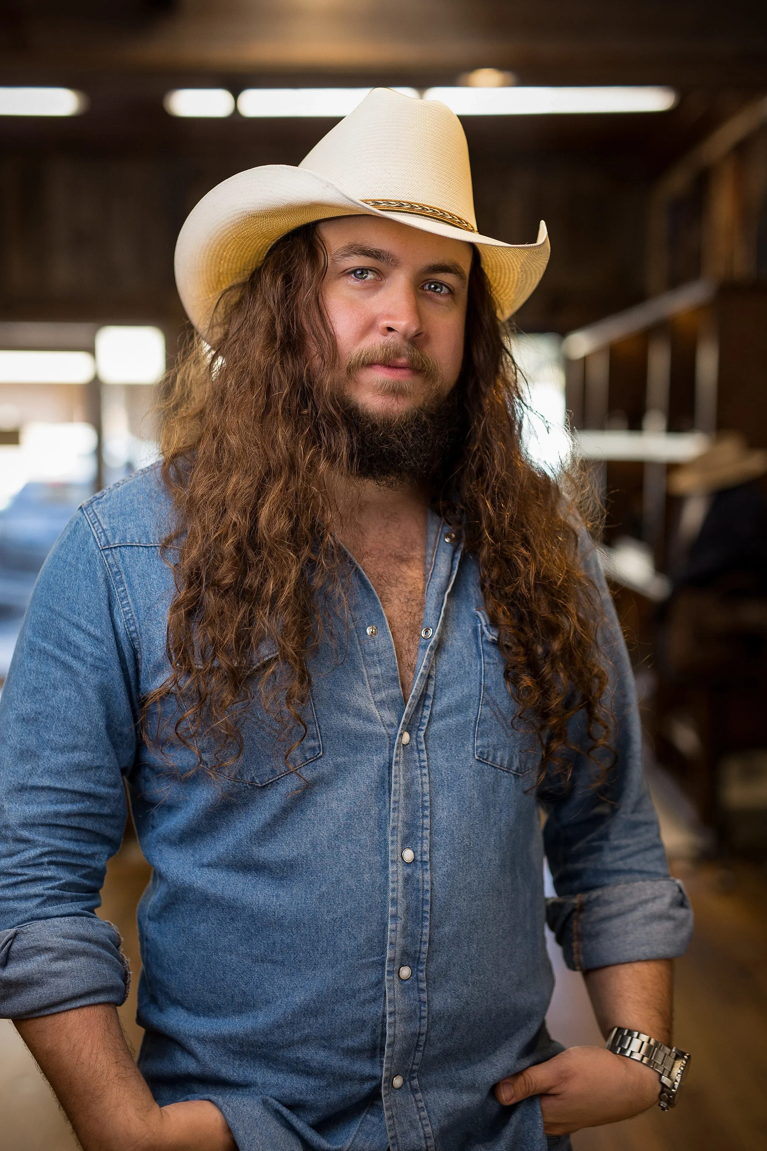 Authentic lifestyle branding portrait of a man with long hair wearing a cowboy hat and denim shirt indoors.