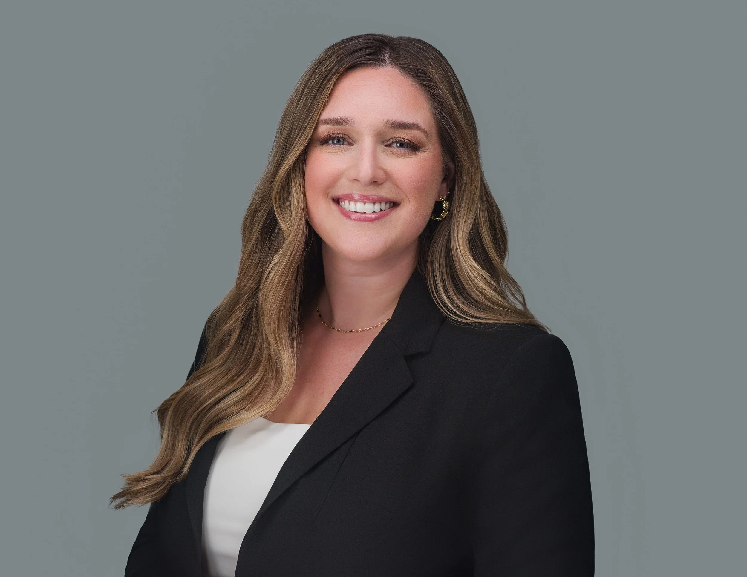 Professional headshot of a woman with long brown hair wearing a black blazer, smiling against a gray background.