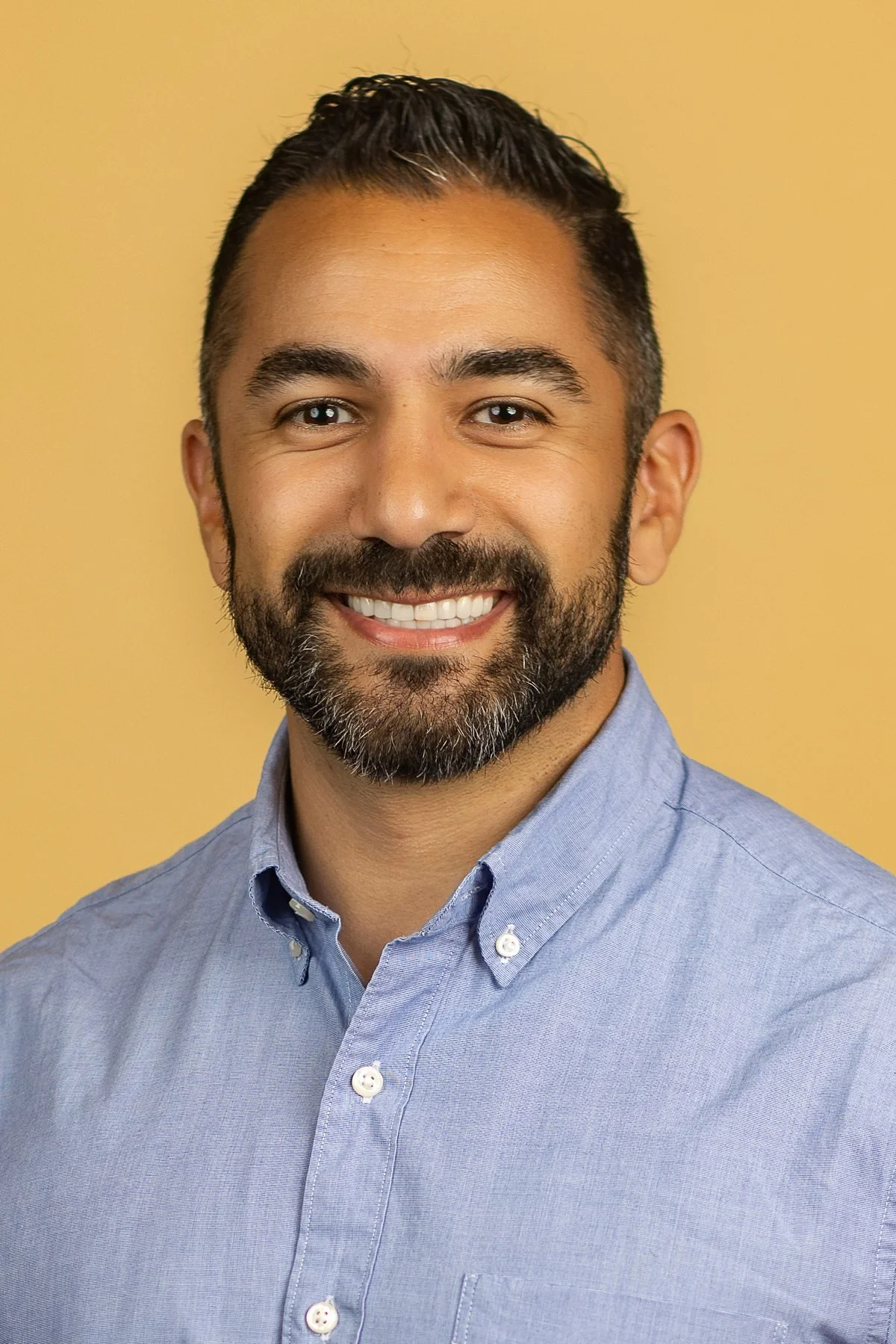 Professional headshot of a man in a light blue shirt with a warm smile on a golden background in San Antonio.
