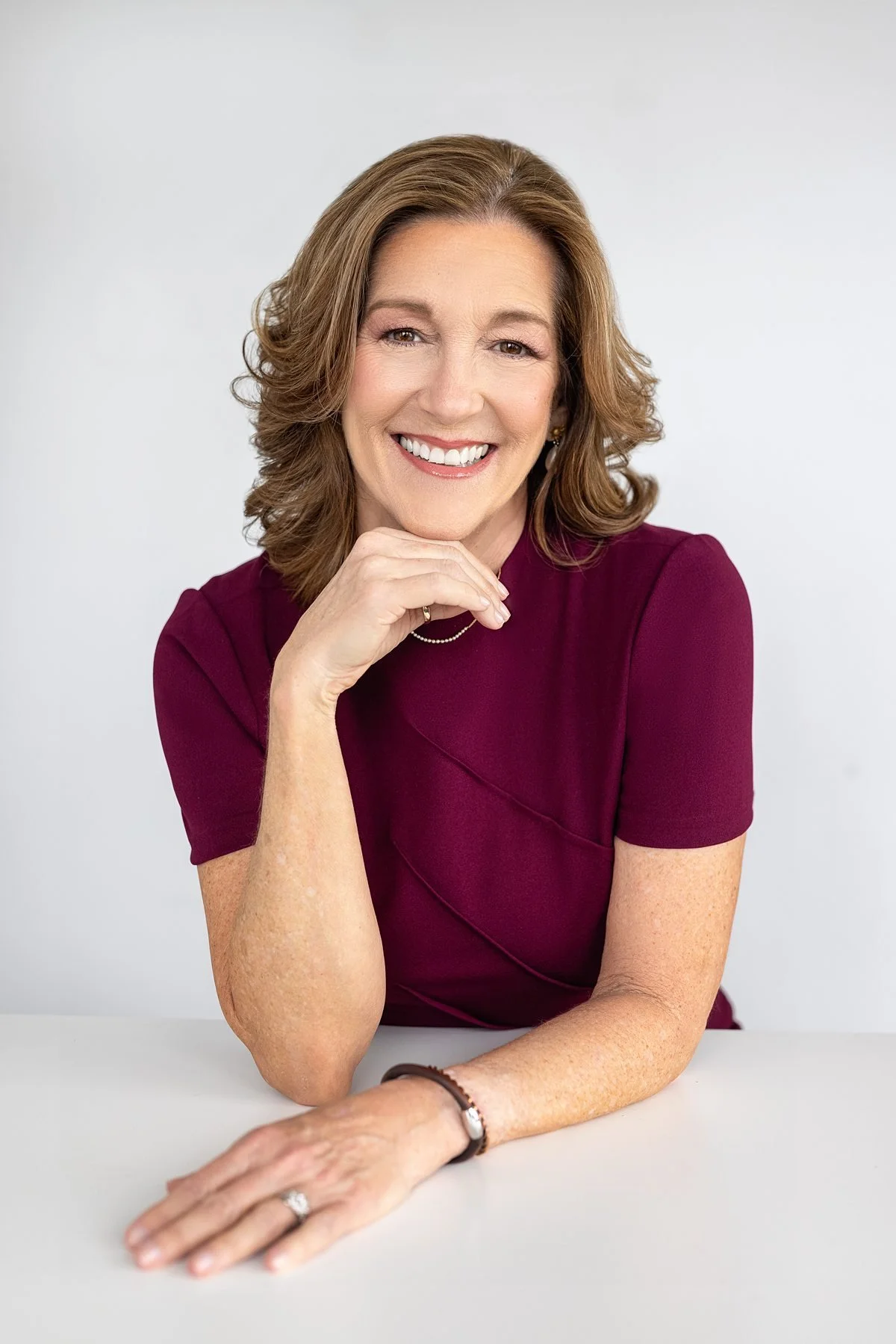 Professional headshot of a woman with brown hair in a burgundy blouse, smiling with hand resting on chin.