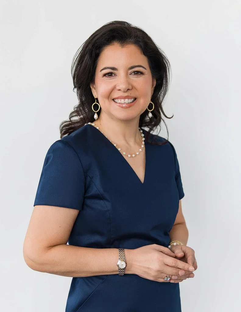 Professional headshot of a woman in a navy blue dress with pearl jewelry, smiling against a clean white background