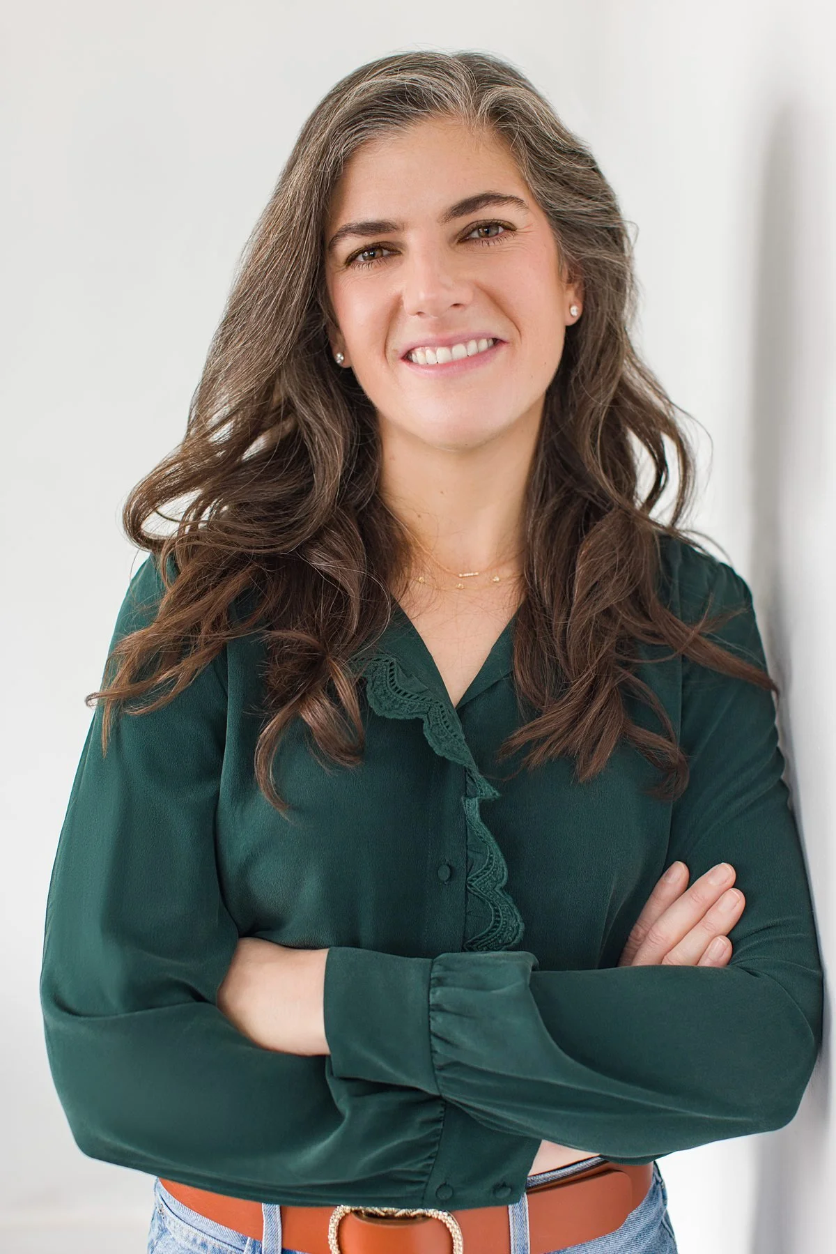 A woman with long wavy brown hair, wearing a dark green blouse with lace details, crossing her arms and smiling at the camera.