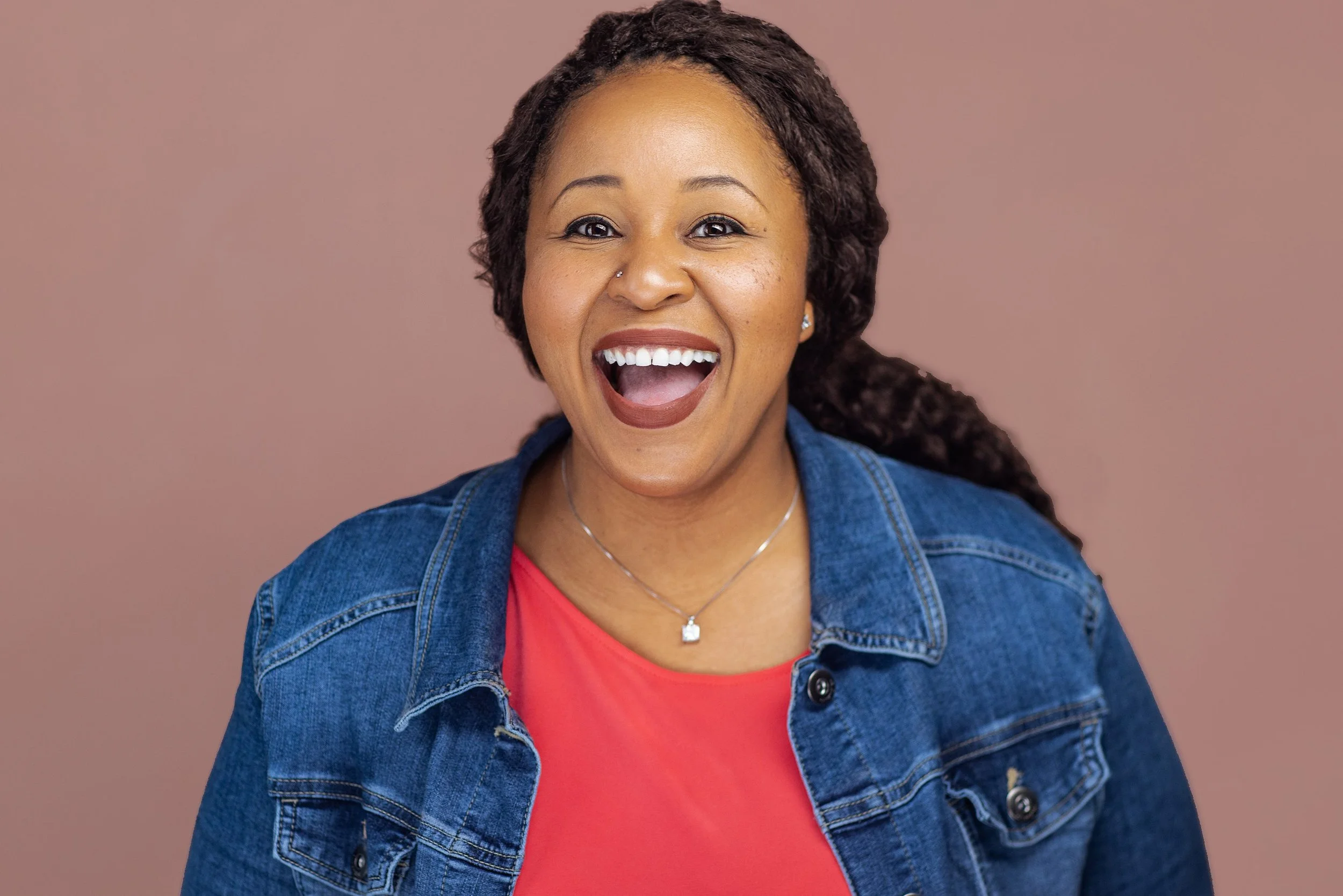 Professional headshot of a woman in a denim jacket and coral top, smiling brightly against a warm background.
