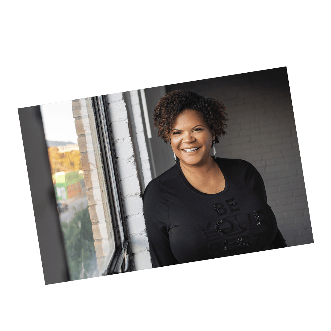 Lifestyle branding headshot of a woman in a black shirt with “Be Your Best” text, smiling by a large window in natural light.
