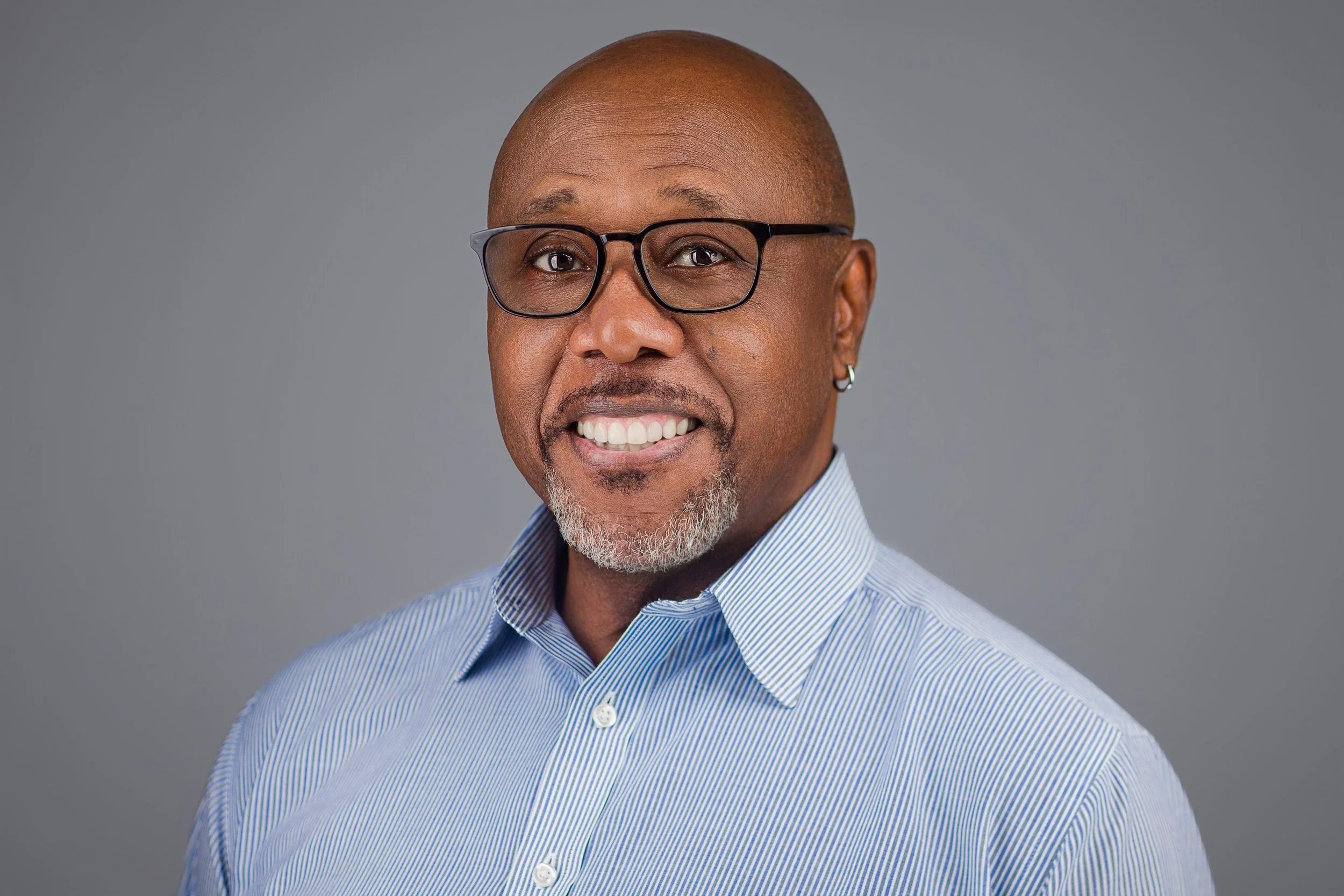 Corporate headshot of a professional man in glasses wearing a blue shirt against a neutral background.