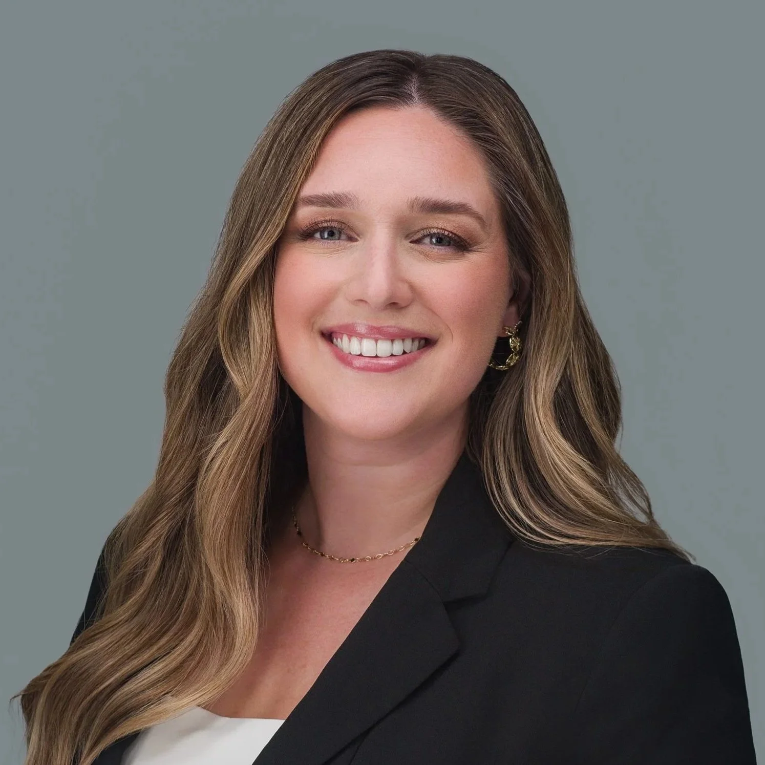 Professional headshot of a woman with long brown hair wearing a black blazer, smiling against a gray background.