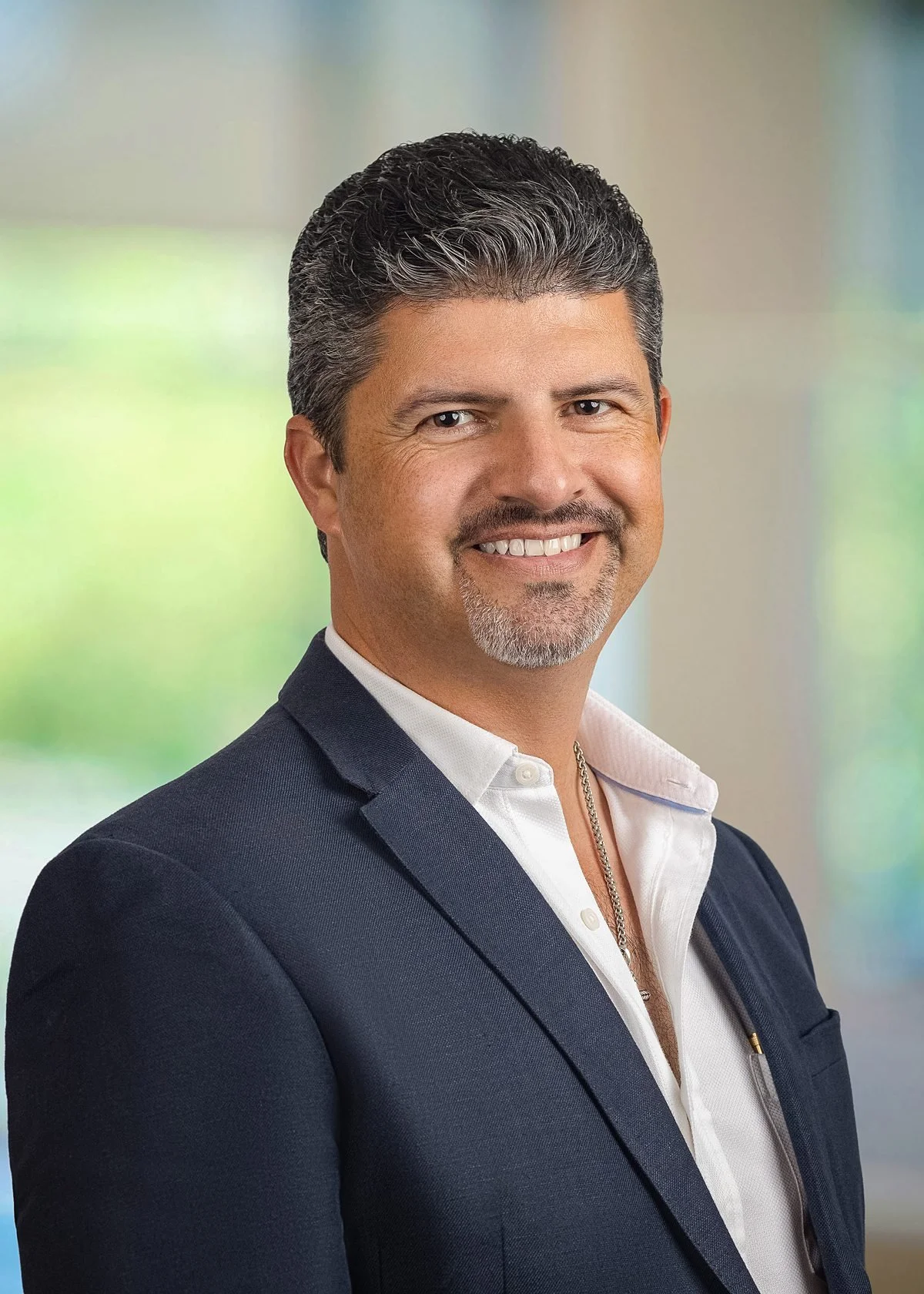 Professional business headshot of a man with salt-and-pepper hair and goatee in a navy blazer and white shirt, smiling against a softly blurred office background.
