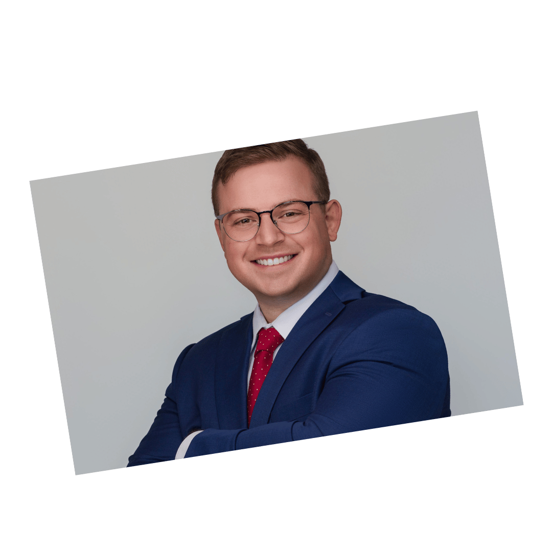Professional headshot of a man in a blue suit with a red tie and glasses, smiling in front of a light background.