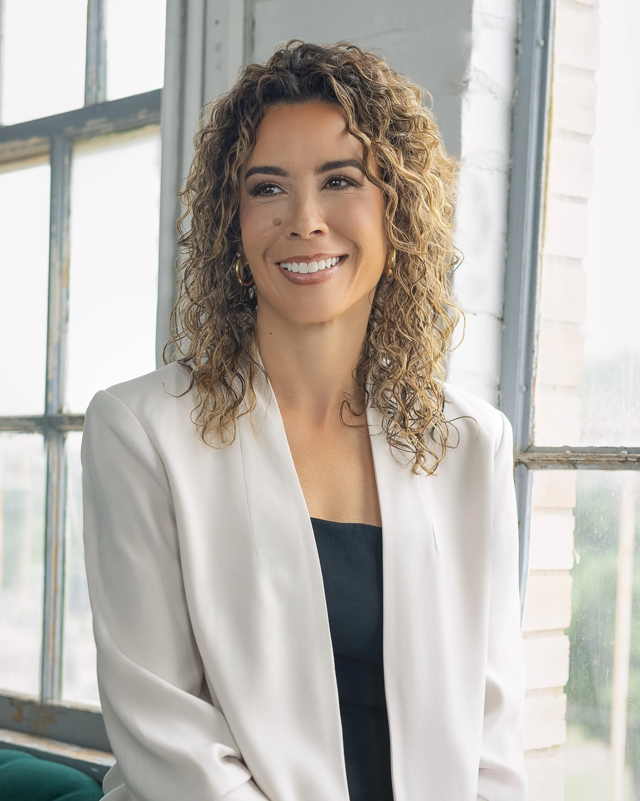 Professional headshot of a woman with curly hair wearing a white blazer, smiling by a window, San Antonio corporate photography.