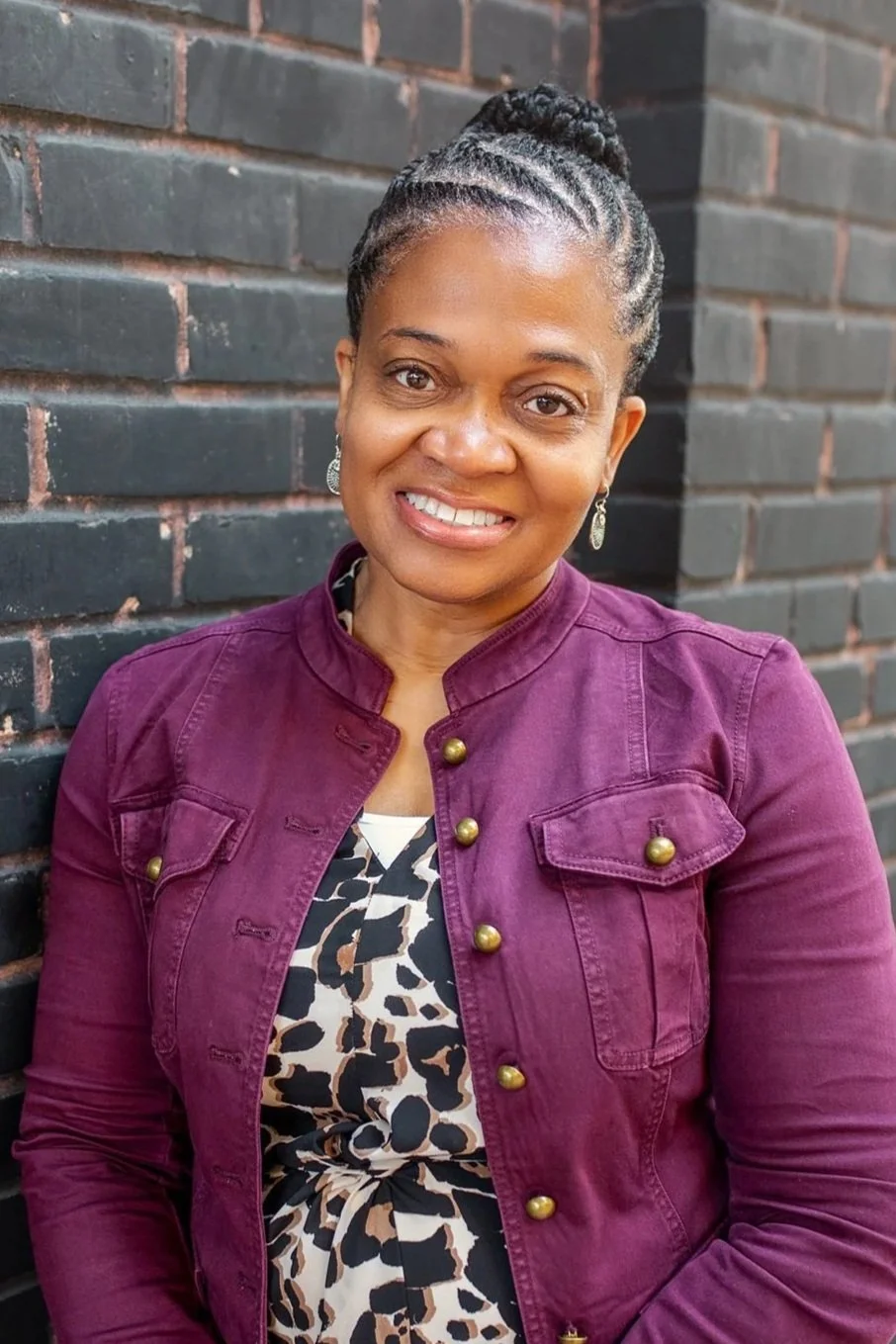 Business portrait of a woman with braided hair, wearing a purple jacket and patterned blouse, standing against a brick wall.