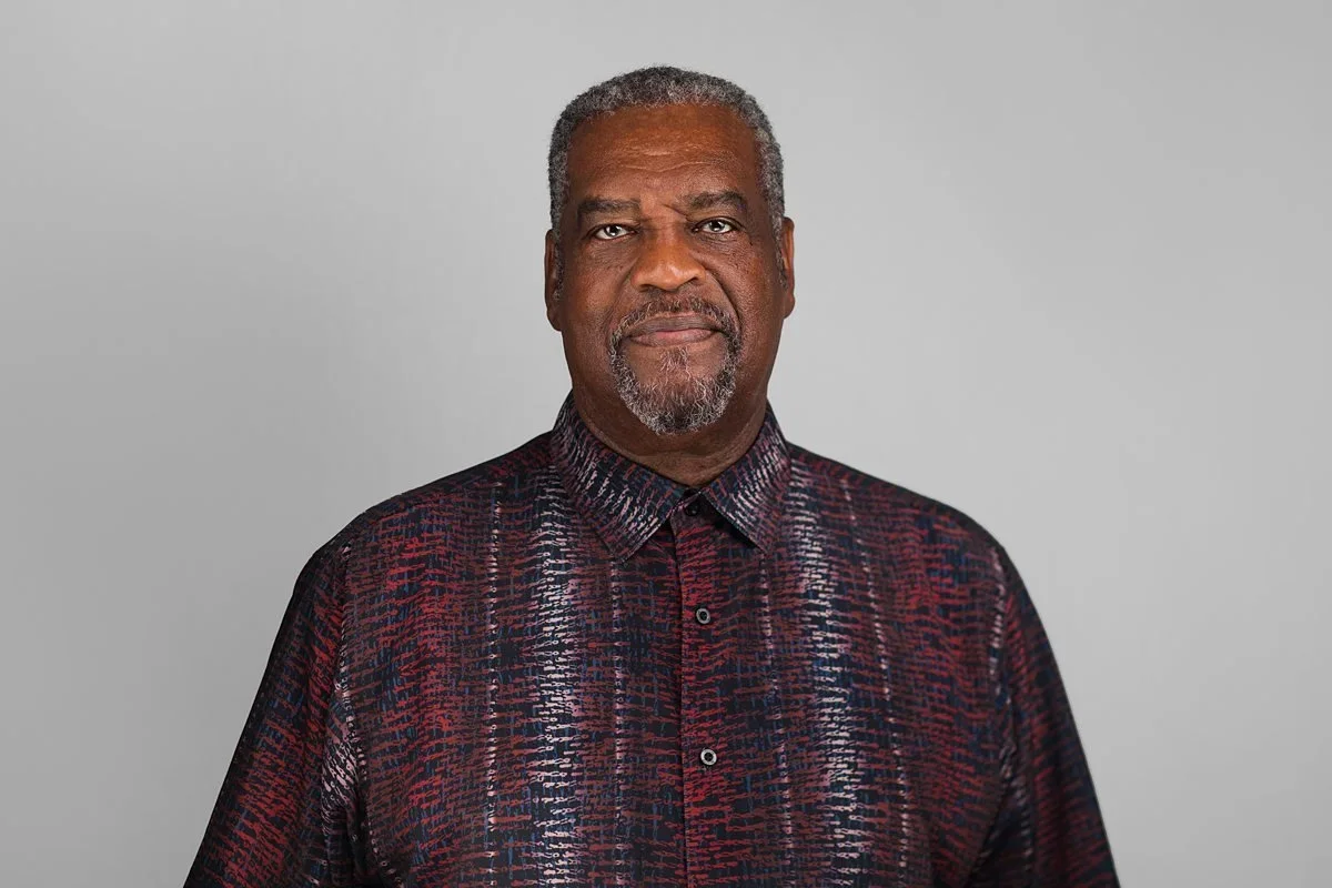Studio headshot of an older man in a patterned red and black shirt on a gray background.