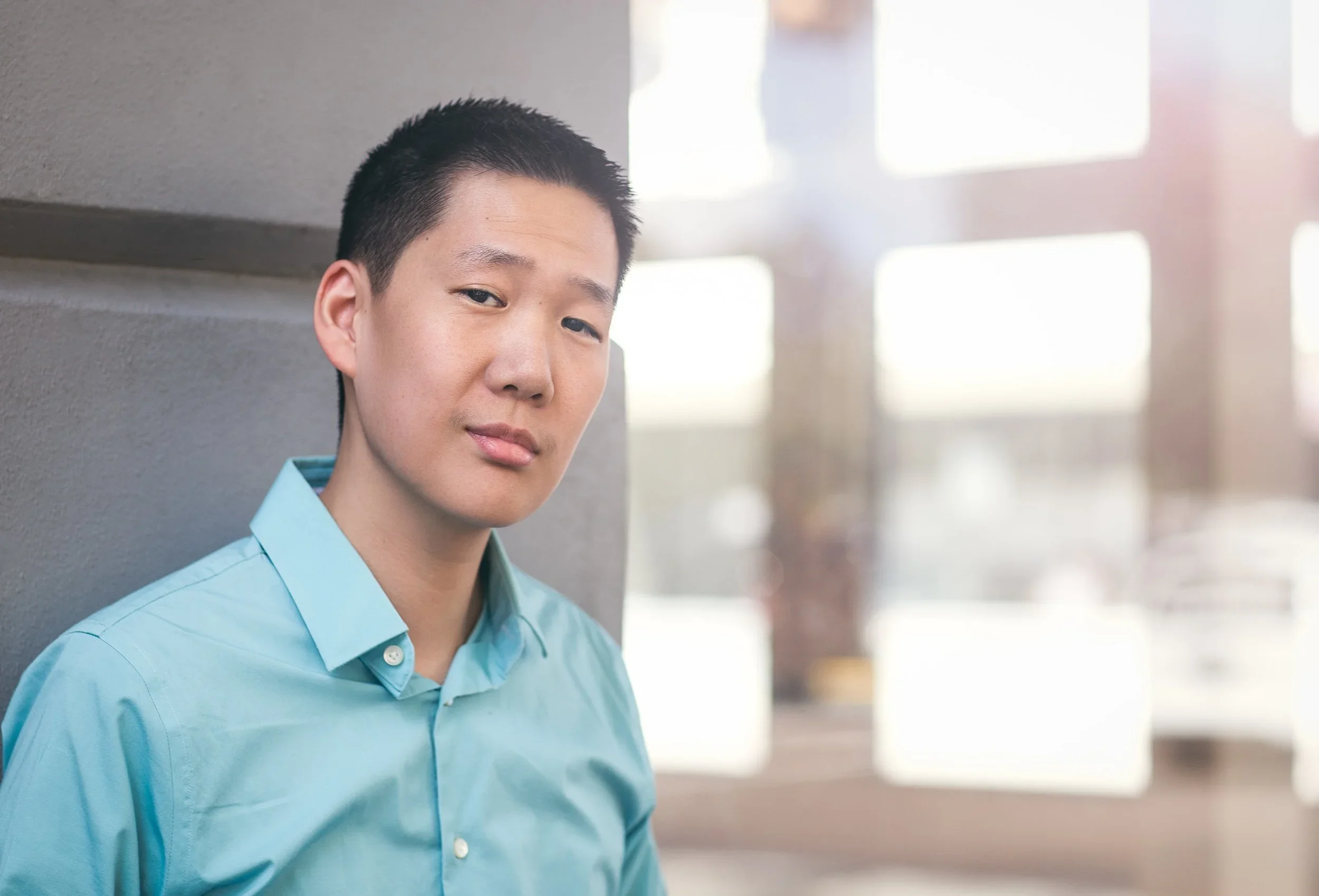 Professional headshot of a man in a teal shirt leaning against a wall with natural light and blurred cityscape background.