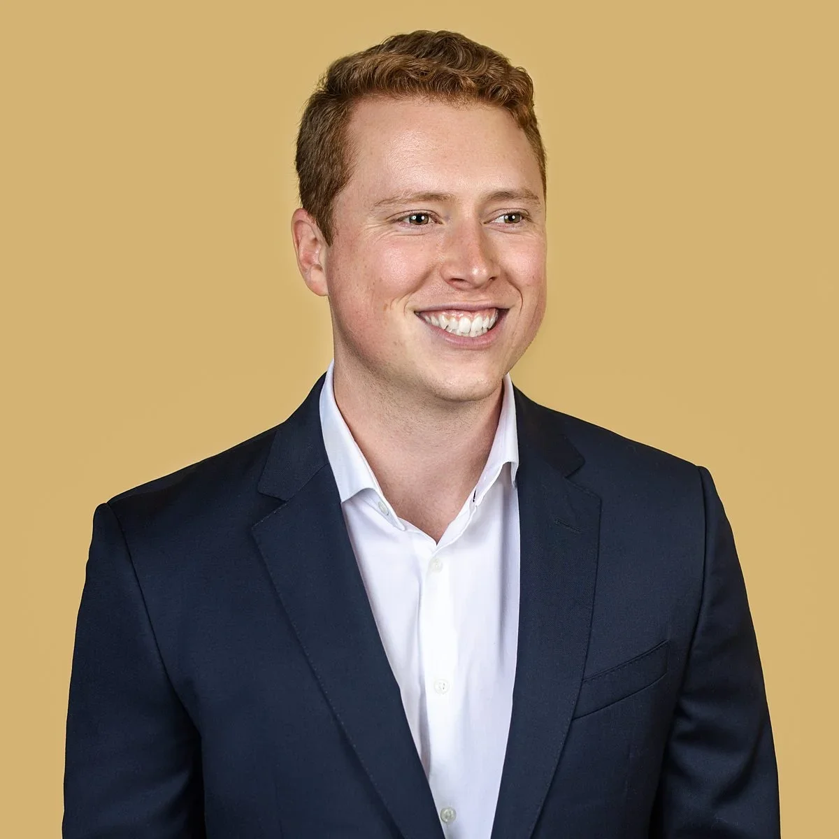 Modern corporate headshot of a young professional in a navy suit against a golden backdrop.