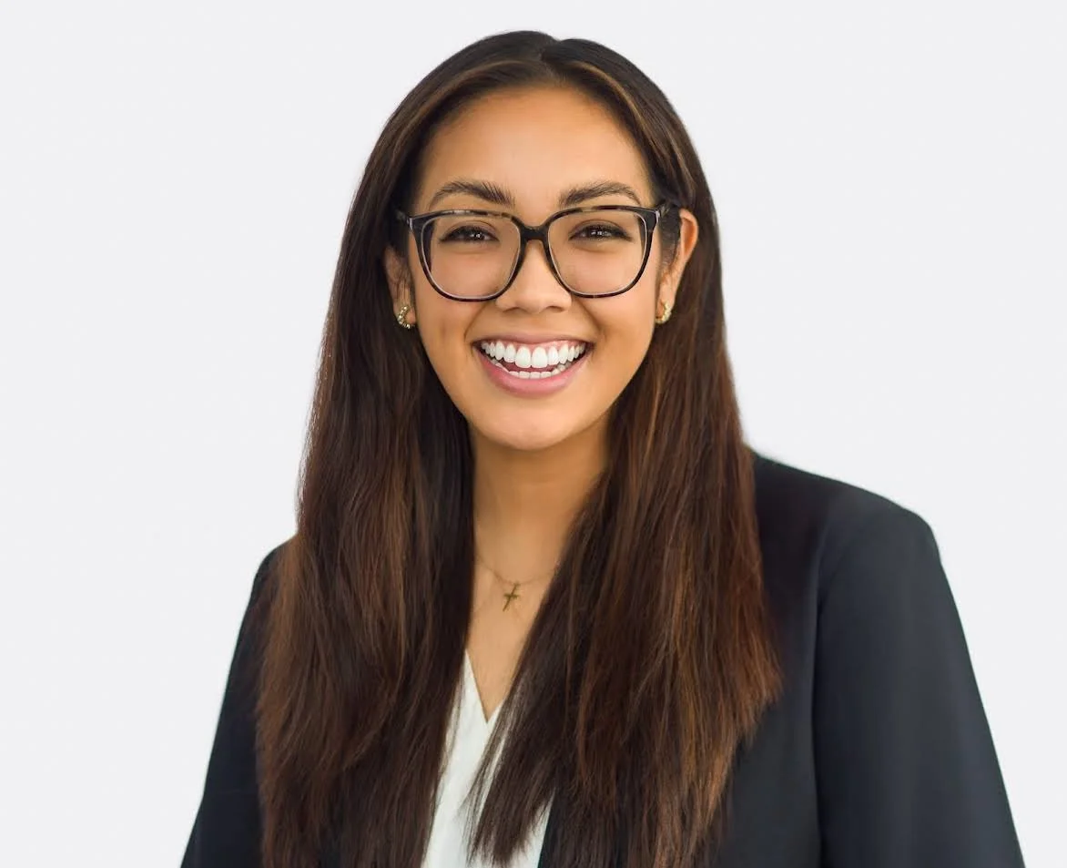 Professional headshot of a young woman with long brown hair and glasses, smiling in a business suit against a white background.