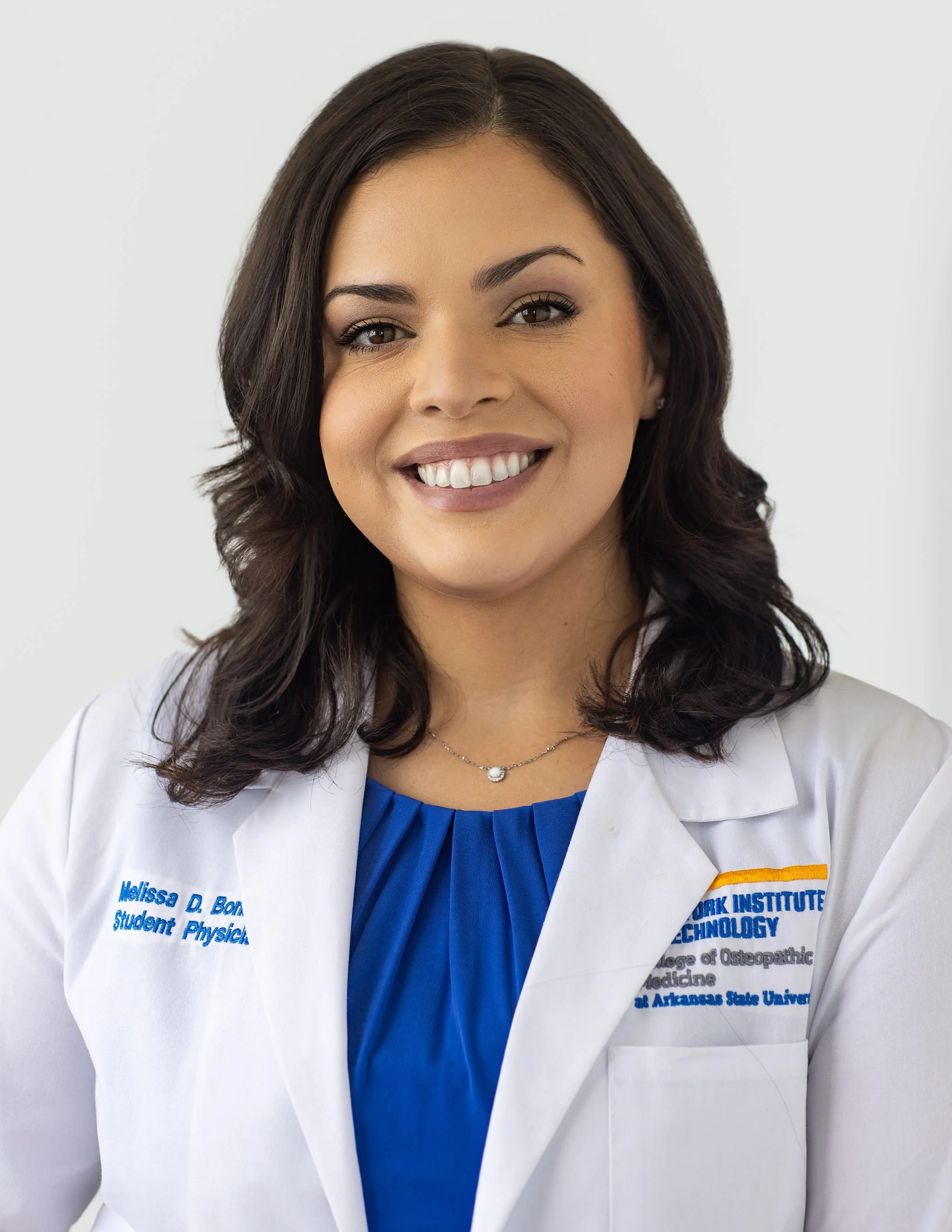 Professional headshot of a female medical student wearing a white coat and blue blouse, smiling confidently against a light background.