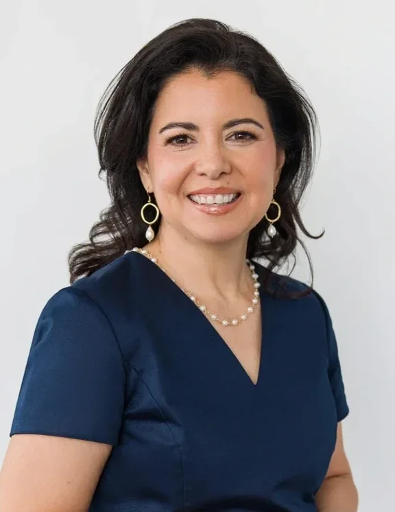 Professional headshot of a woman in a navy blue dress with pearl jewelry, smiling against a clean white background.