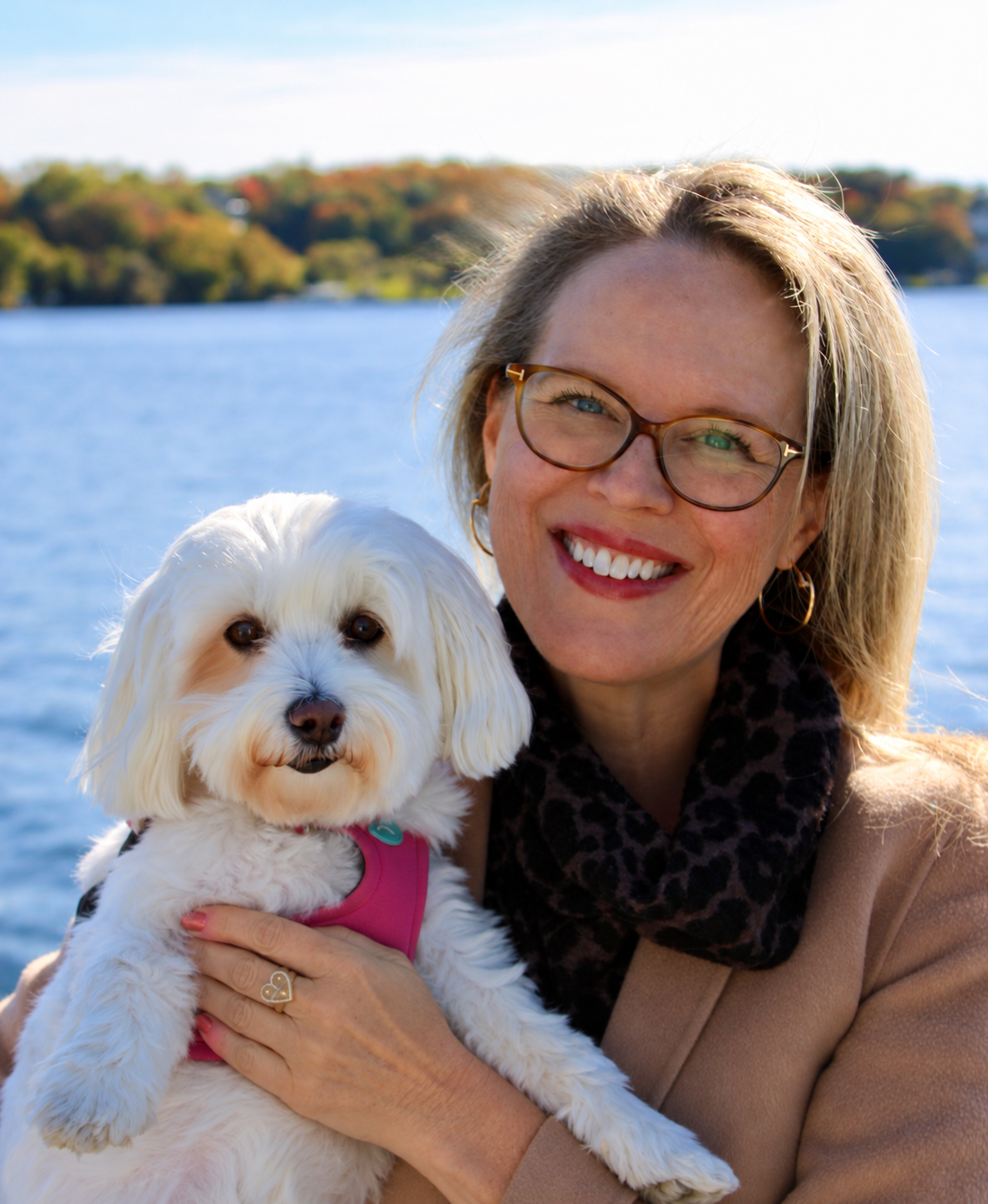 A woman with blonde hair cuddling Maltese dog by Lake Geneva