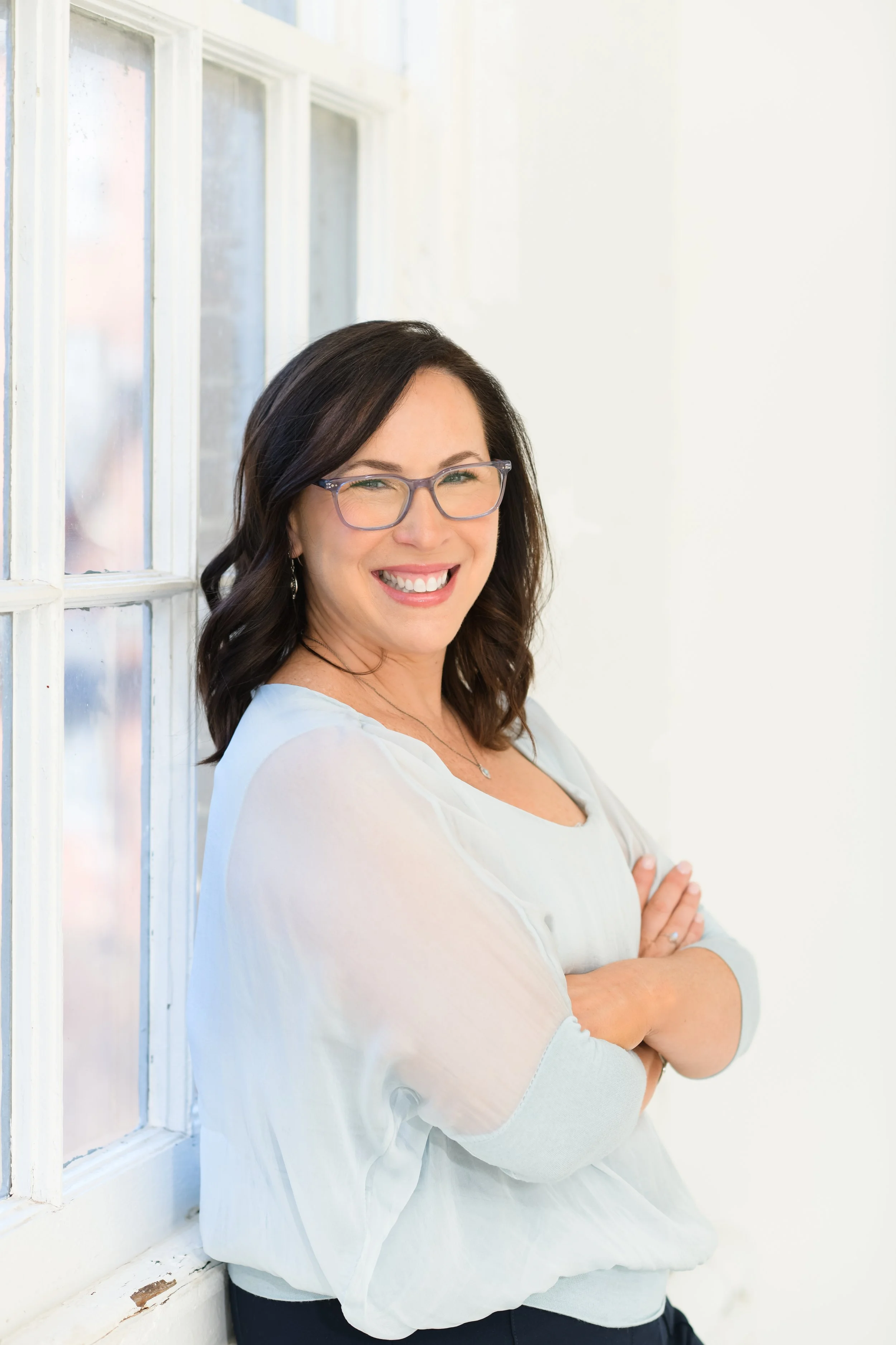Jill Vetstein is standing in her Natick MA Therapy office leaning against a window. She's wearing glasses and a flowy top.