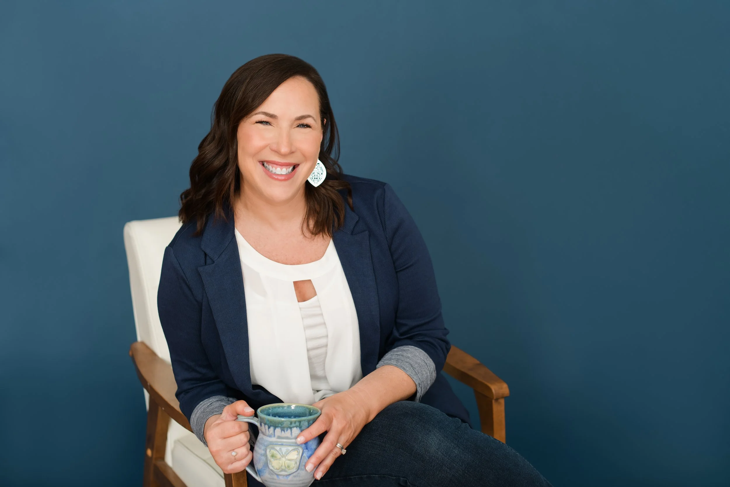 Jill is seated in a chair, smiling and holding a handmade clay mug with a butterfly on the front.