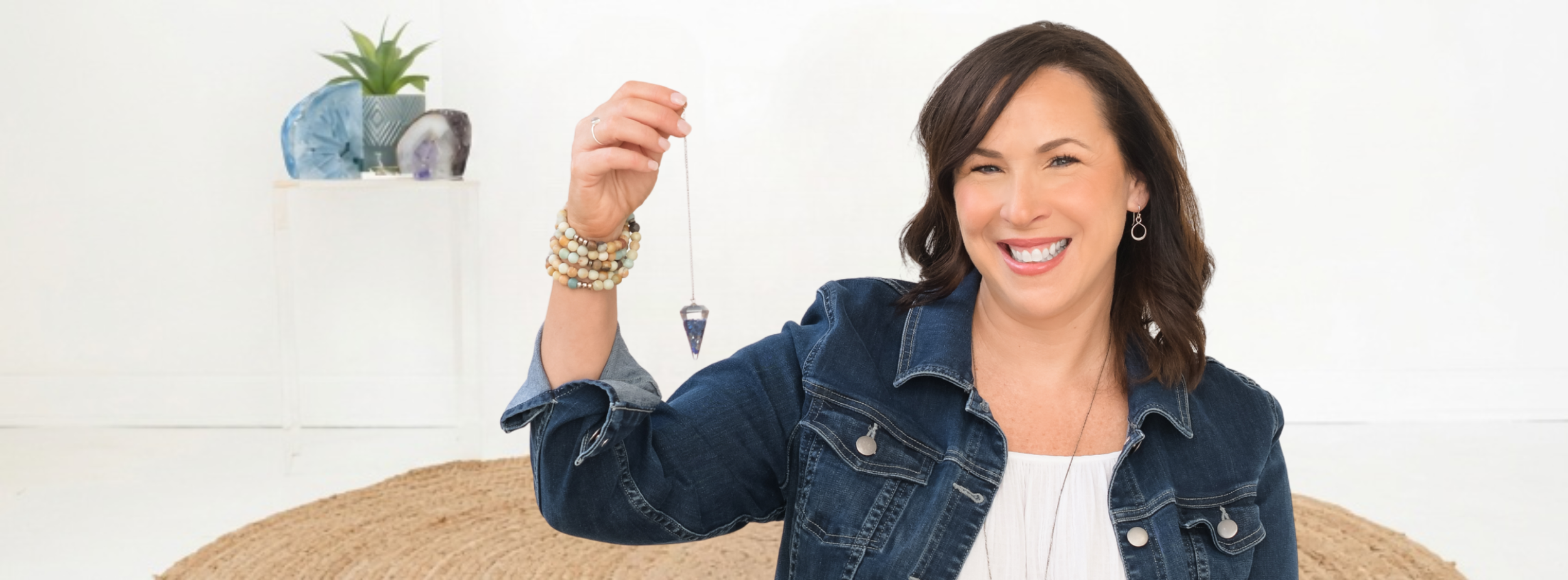 Jill Vetstein, spiritual coach, is sitting on the floor holding a pendulum and smiling. There are crystals and a plant in the background.