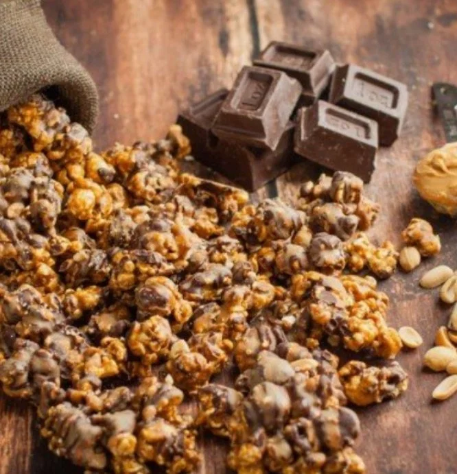 Chocolate drizzled caramel popcorn, chocolate pieces, and peanuts on a wooden surface.