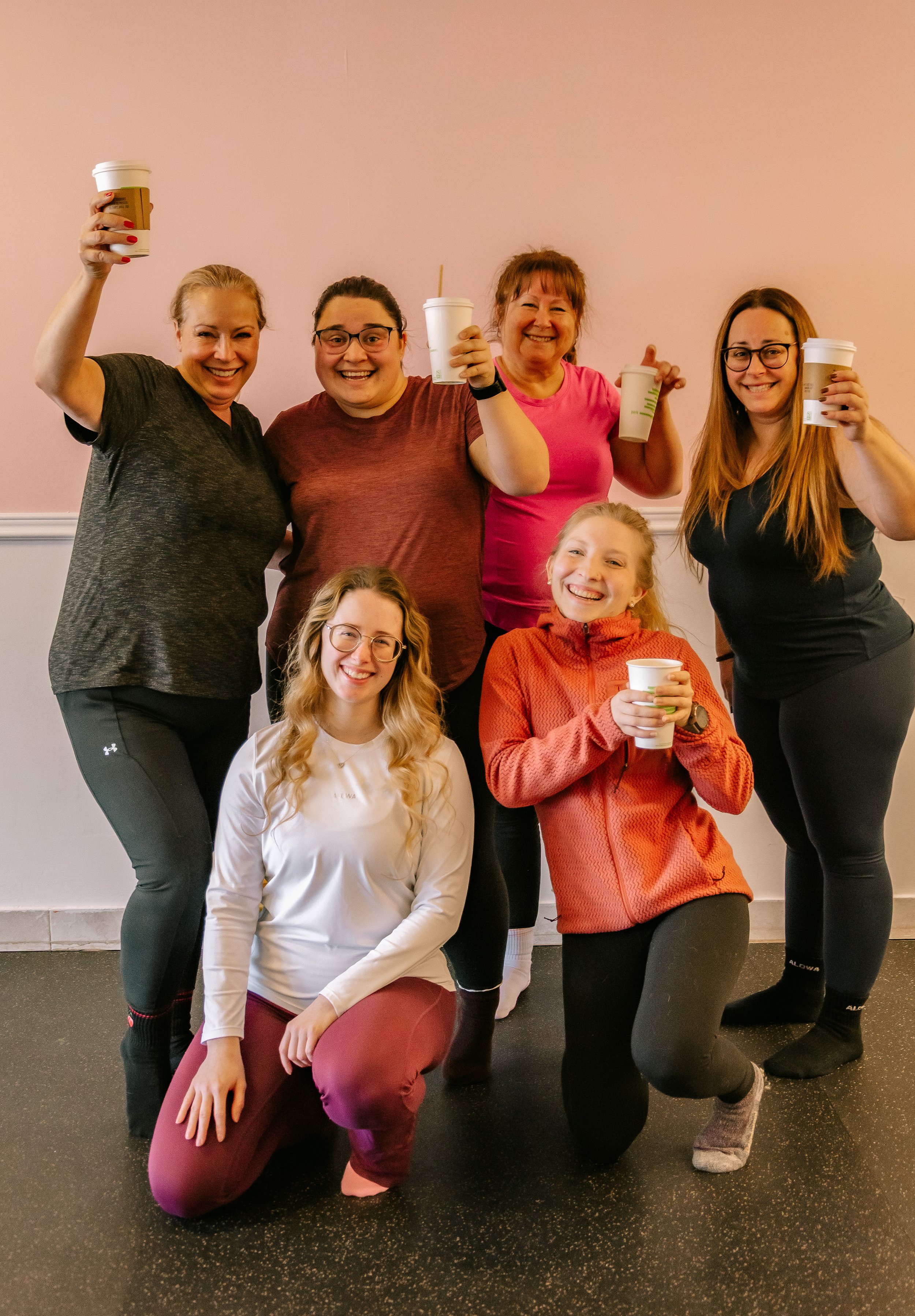 Groupe de sept femmes souriantes portant des vêtements de sport, posant dans une salle avec un mur rose clair en arrière-plan, tenant des gobelets à boire.