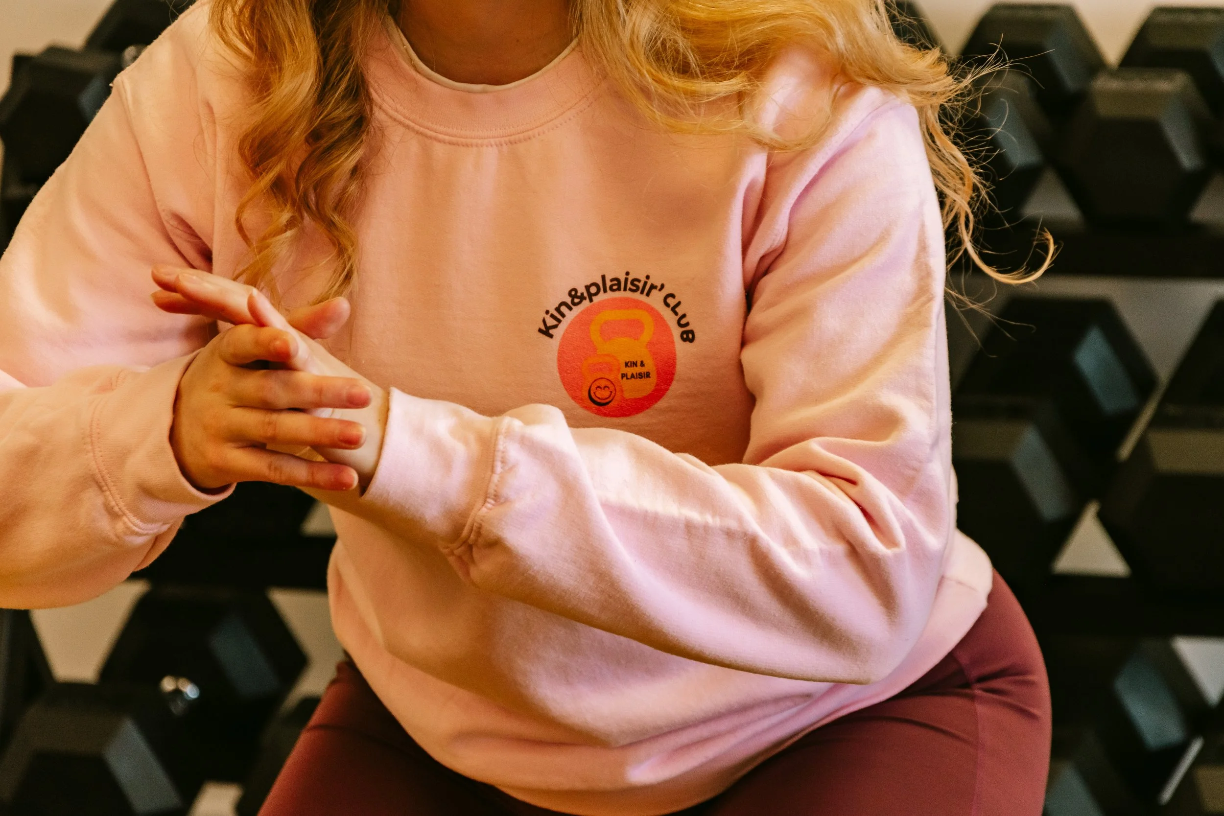 Une personne porte un sweat-shirt rose avec un logo de club de fitness appelé 'Kin & Plaisir'. Elle est assise, les bras croisés et les mains jointes.