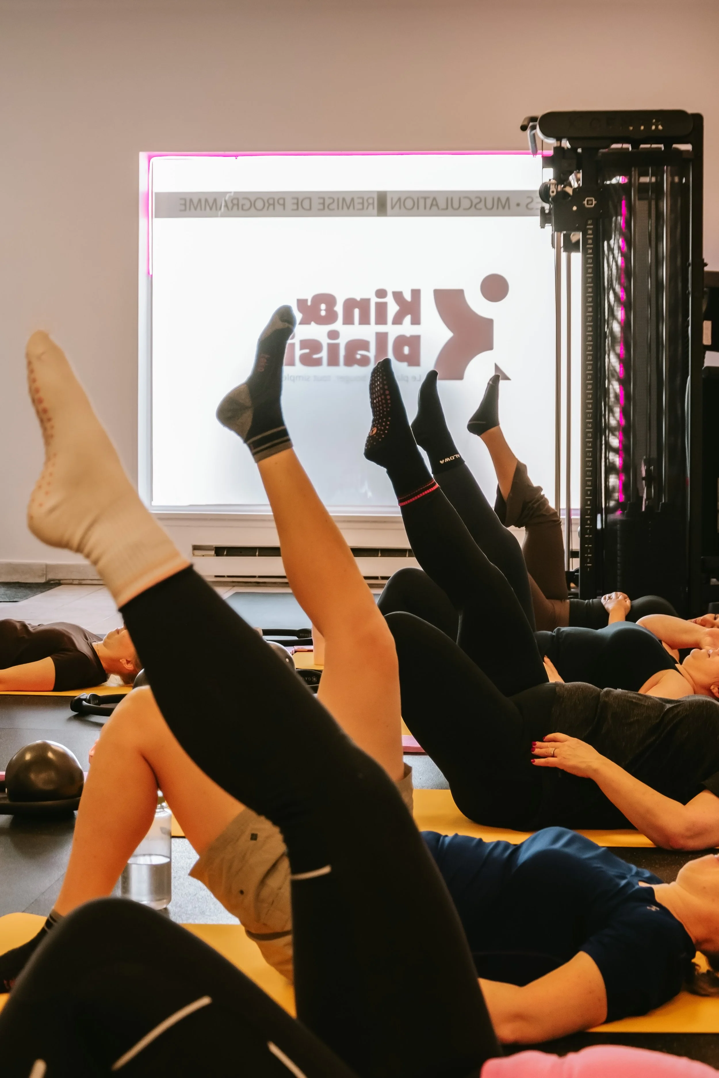 Groupe de personnes faisant des exercices de pilates ou d'étirement sur des tapis en salle de sport, avec une fenêtre derrière elles affichant une publicité pour un studio de remise en forme.