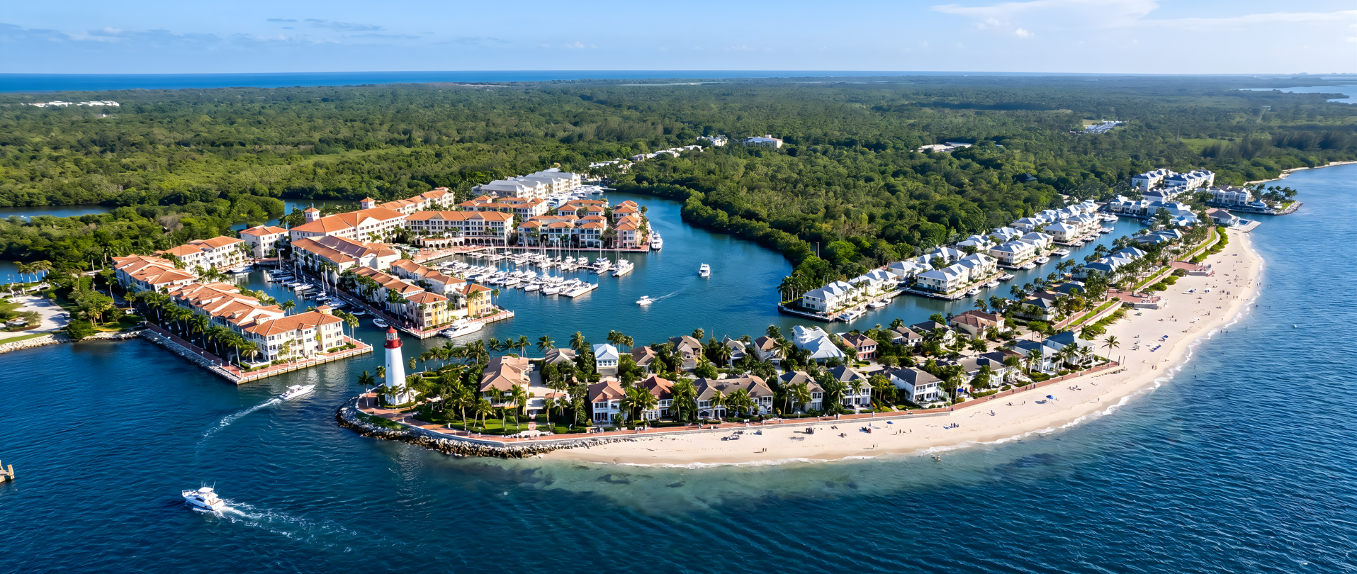 Aerial view of a coastal resort with white villas, a marina with boats, a lighthouse, and a sandy beach with umbrellas and people, surrounded by lush greenery and the ocean.