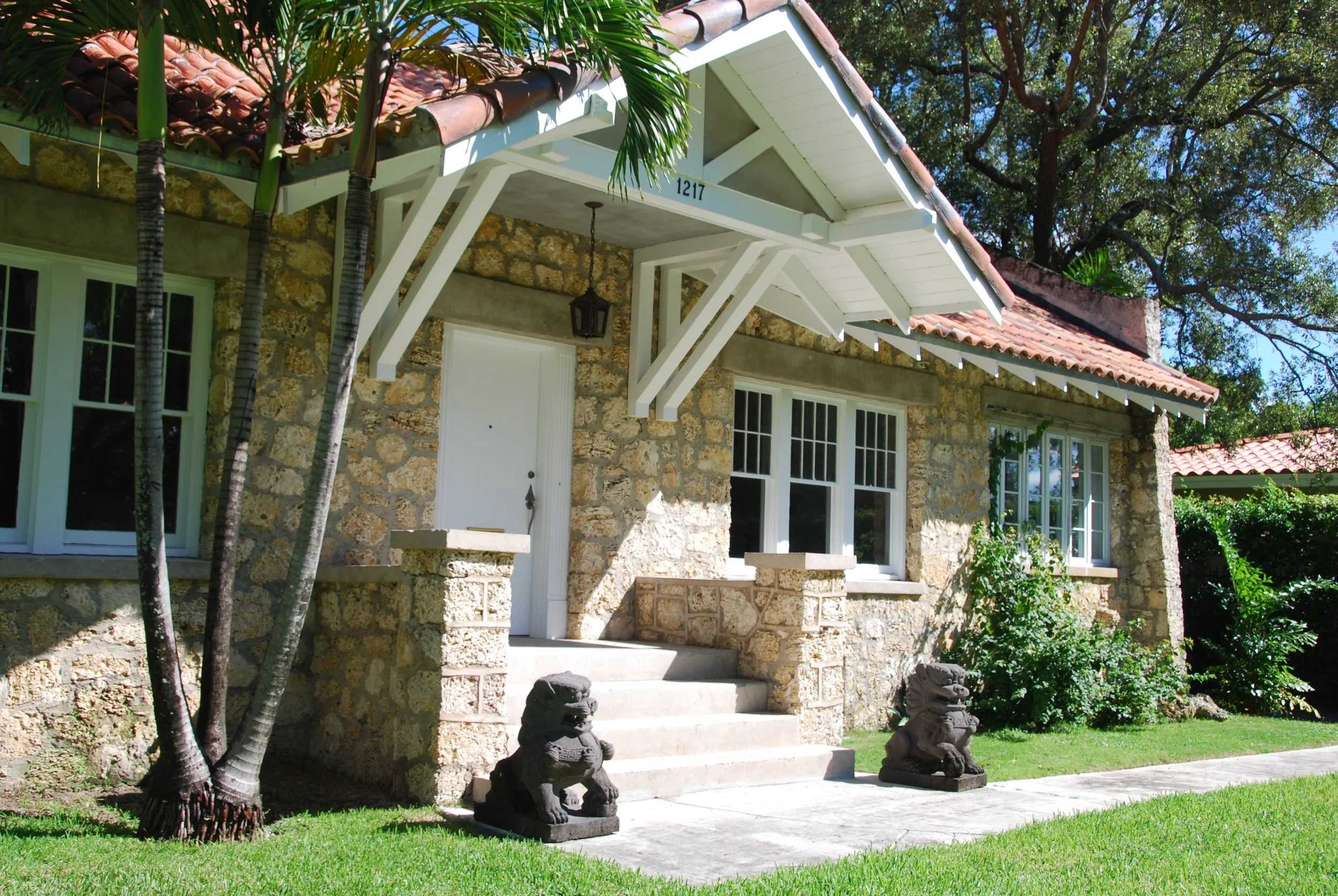 The image shows the front of a stone house with a tiled roof and white trim. There are steps leading to a white door with black lion statues on each side. Two tiki statues are on the ground near the steps. Tall trees and bright green grass surround t