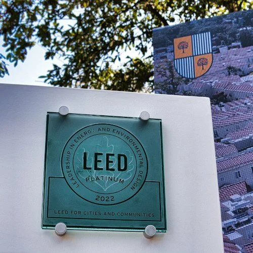 Glass plaque with LEED Platinum 2022 certification for leadership in energy and environmental design, LEED for cities and communities, mounted on a white board. A shield-shaped flag with a tree and blue stripes is visible in the background outdoors.
