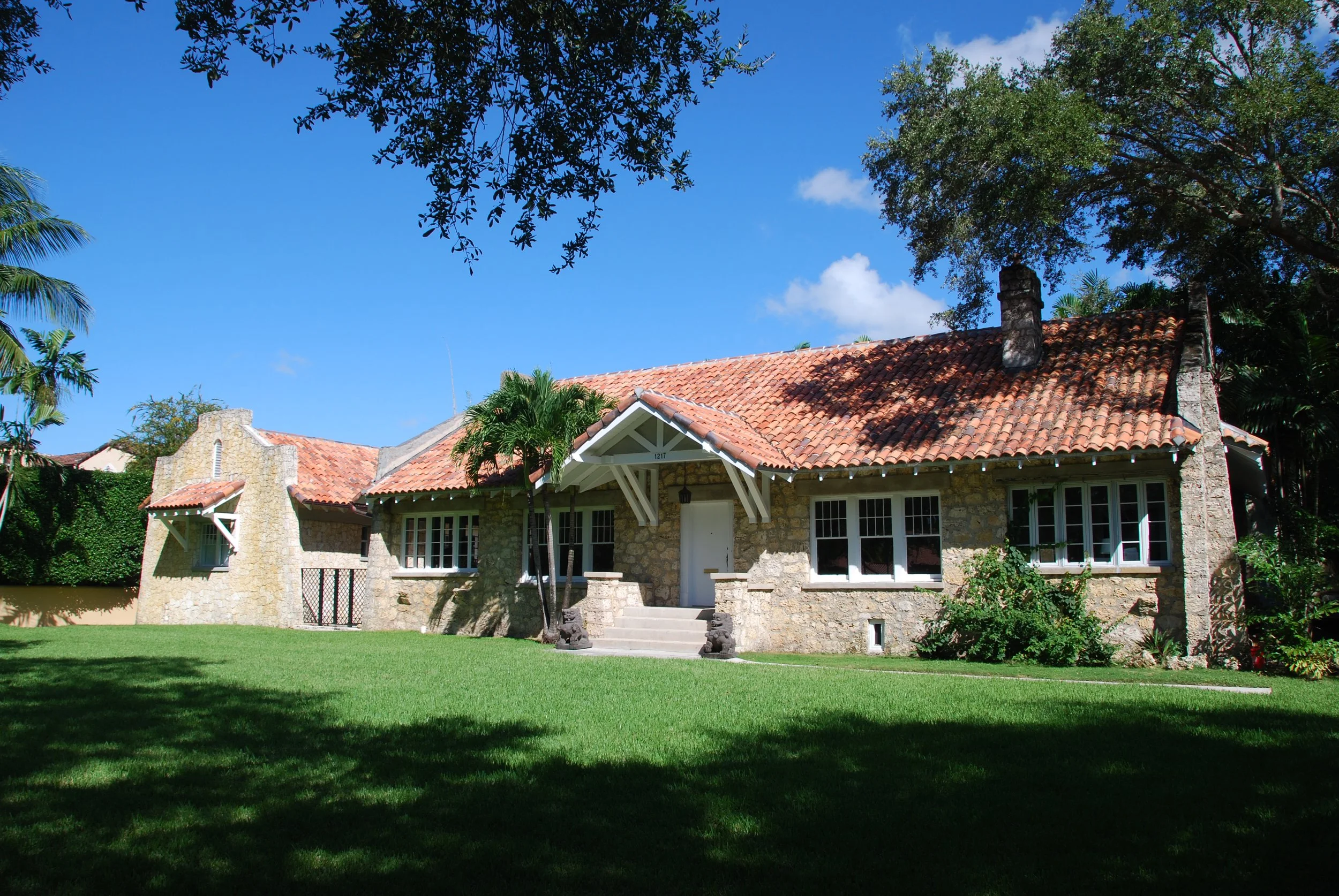 A stone house with a red tile roof, surrounded by lush green lawn and trees under a blue sky.