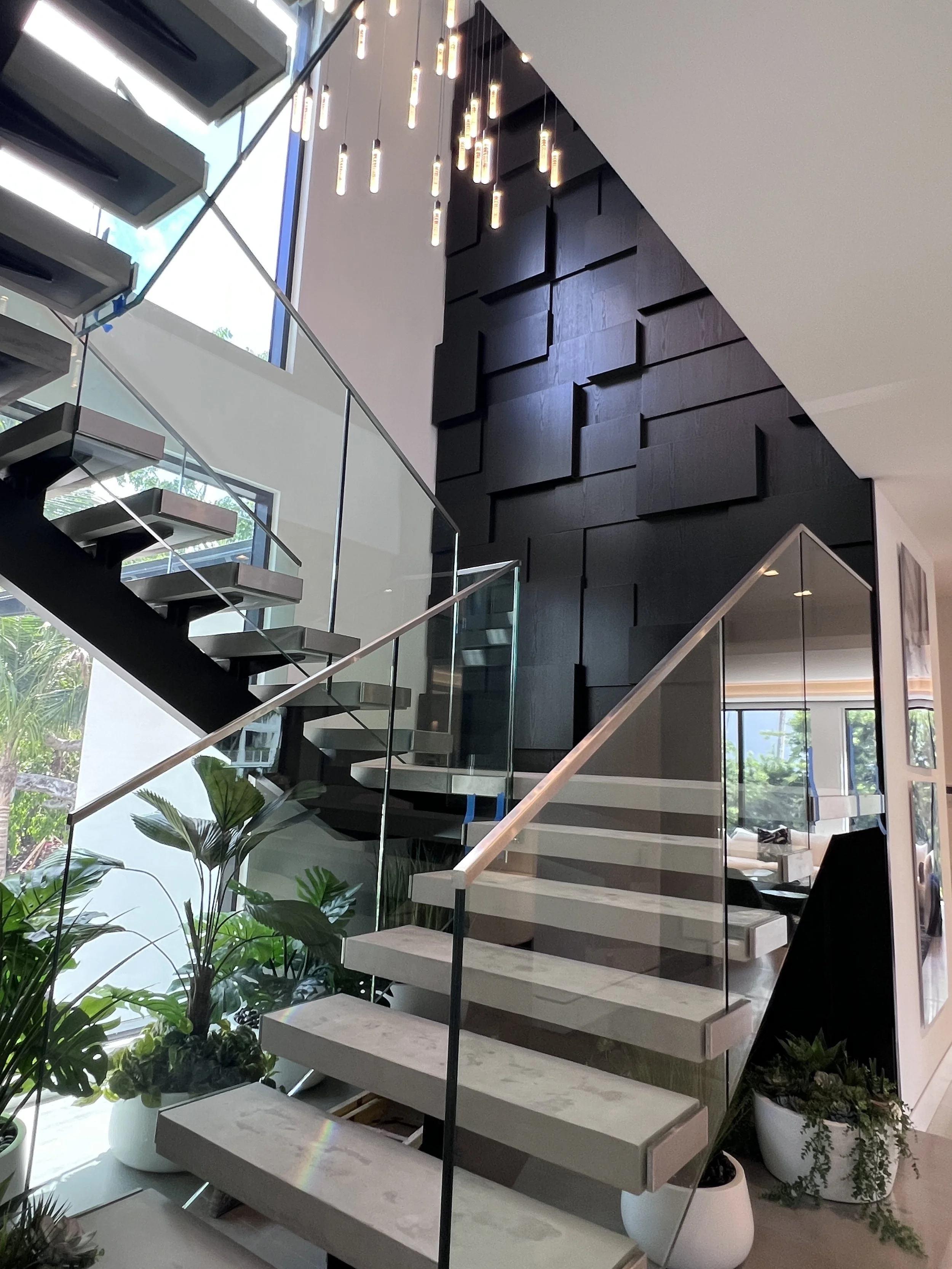 Modern interior staircase with black and white steps, glass railing, black textured wall, decorative pendant lights, and indoor plants near large windows.