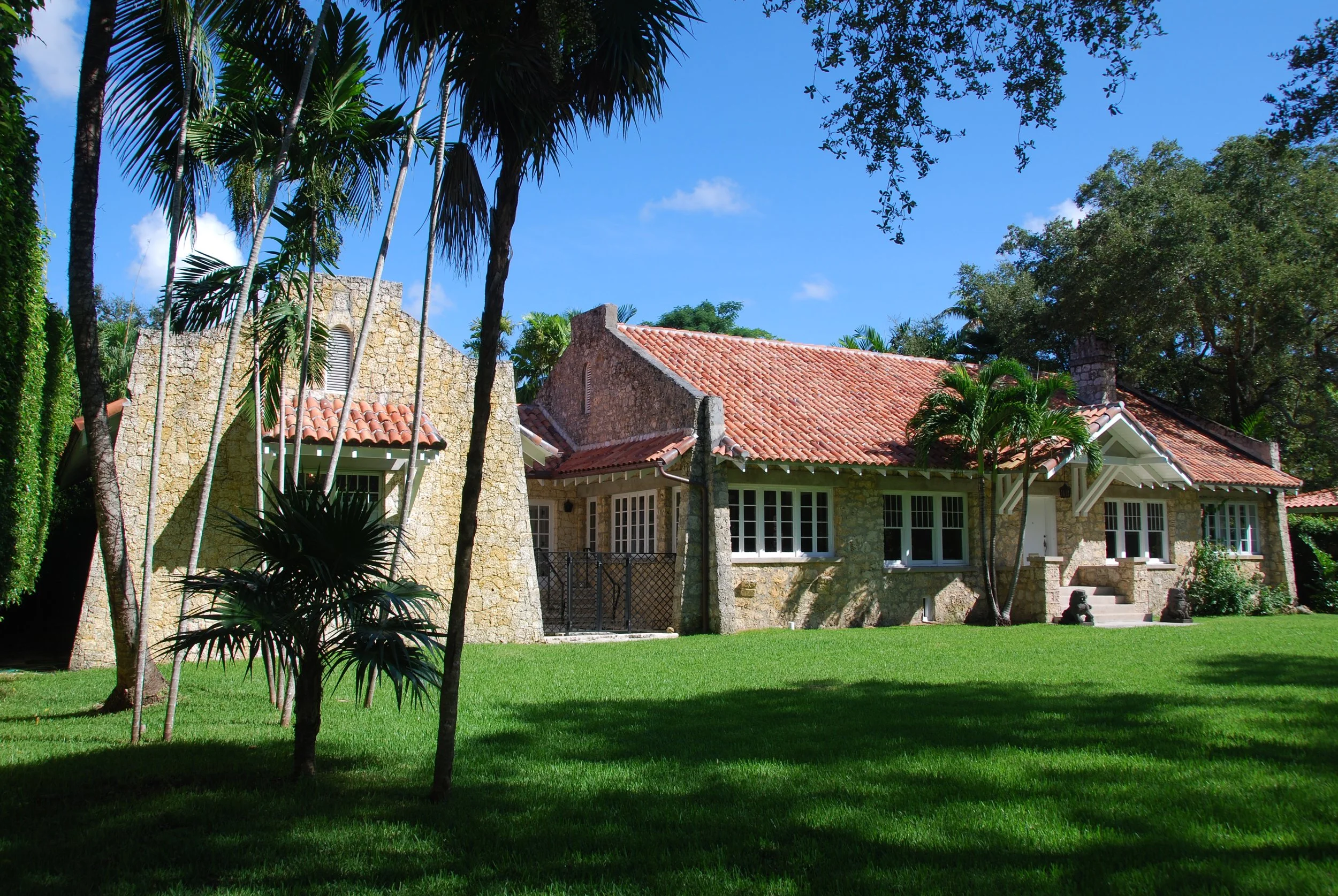 Stone house with red tile roof surrounded by lush green lawn and palm trees under a blue sky.