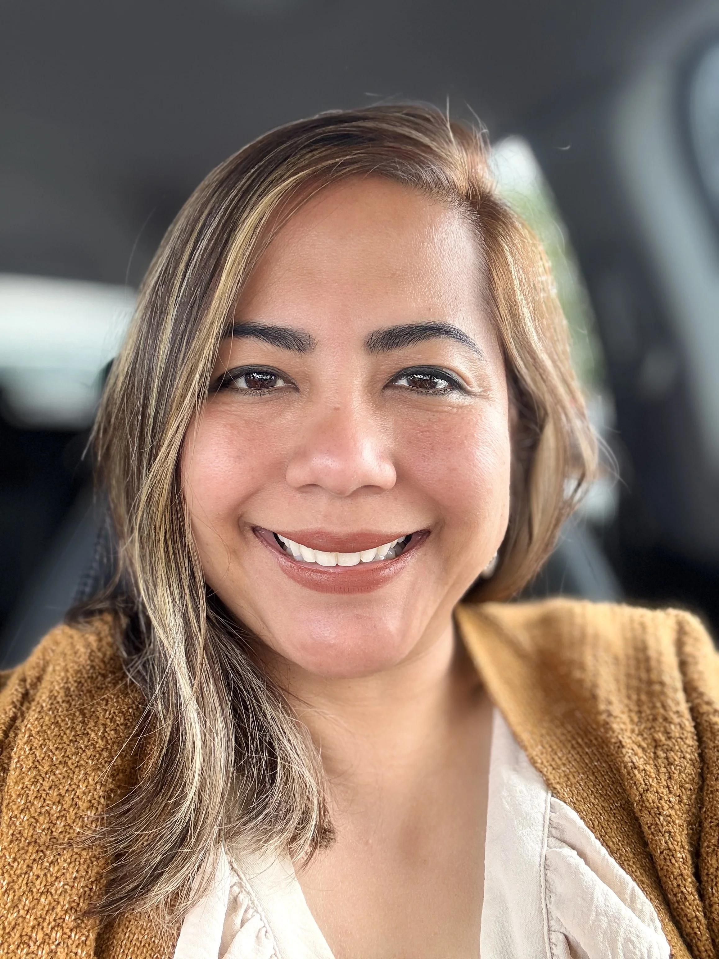 A smiling woman with light brown hair with blonde highlights, wearing a white top and a brown textured jacket, sitting inside a vehicle.