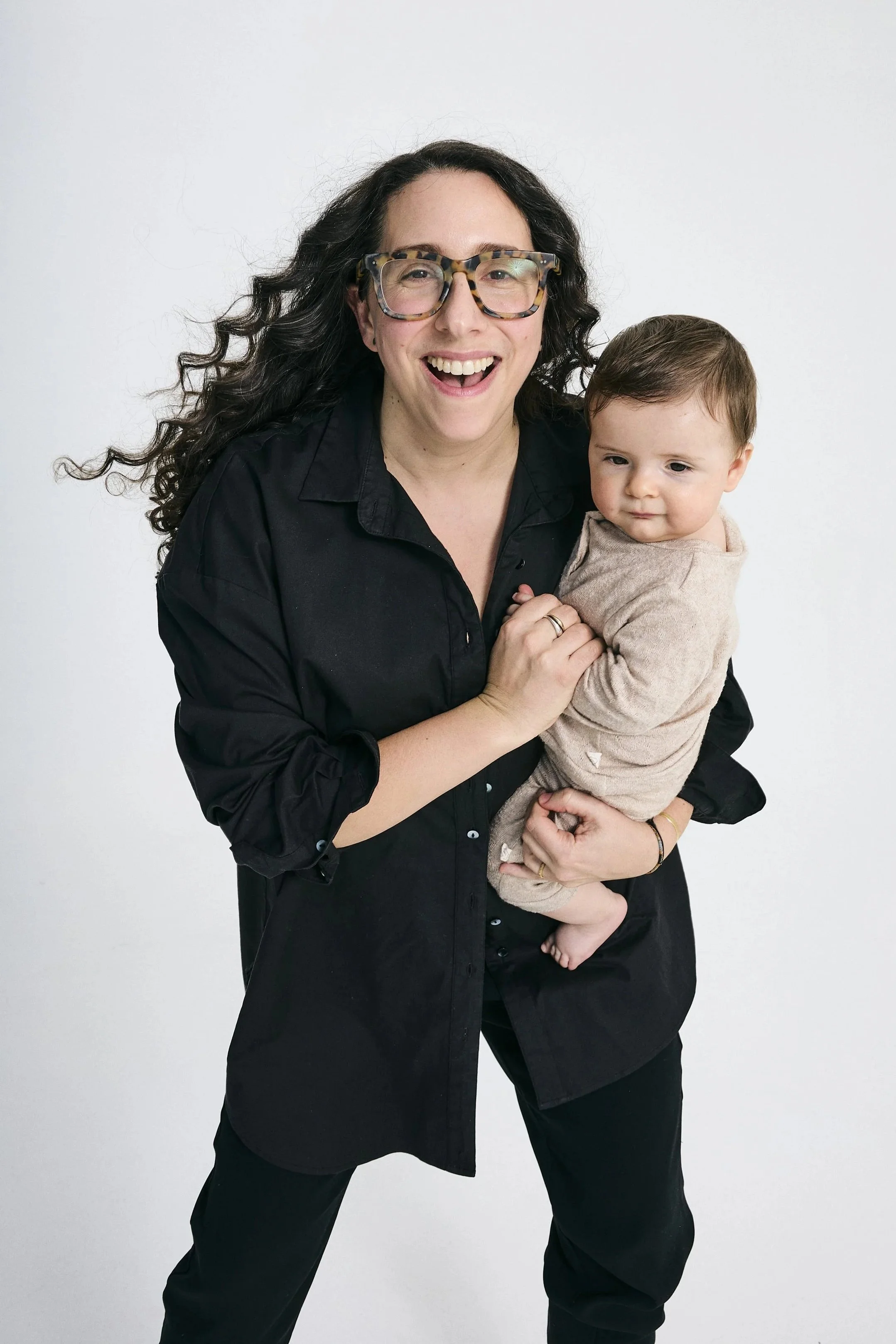 A smiling woman with curly hair and large tortoiseshell glasses is holding a young child with short brown hair and a beige shirt. The woman is wearing a black shirt and pants, and they are posing against a plain white background.