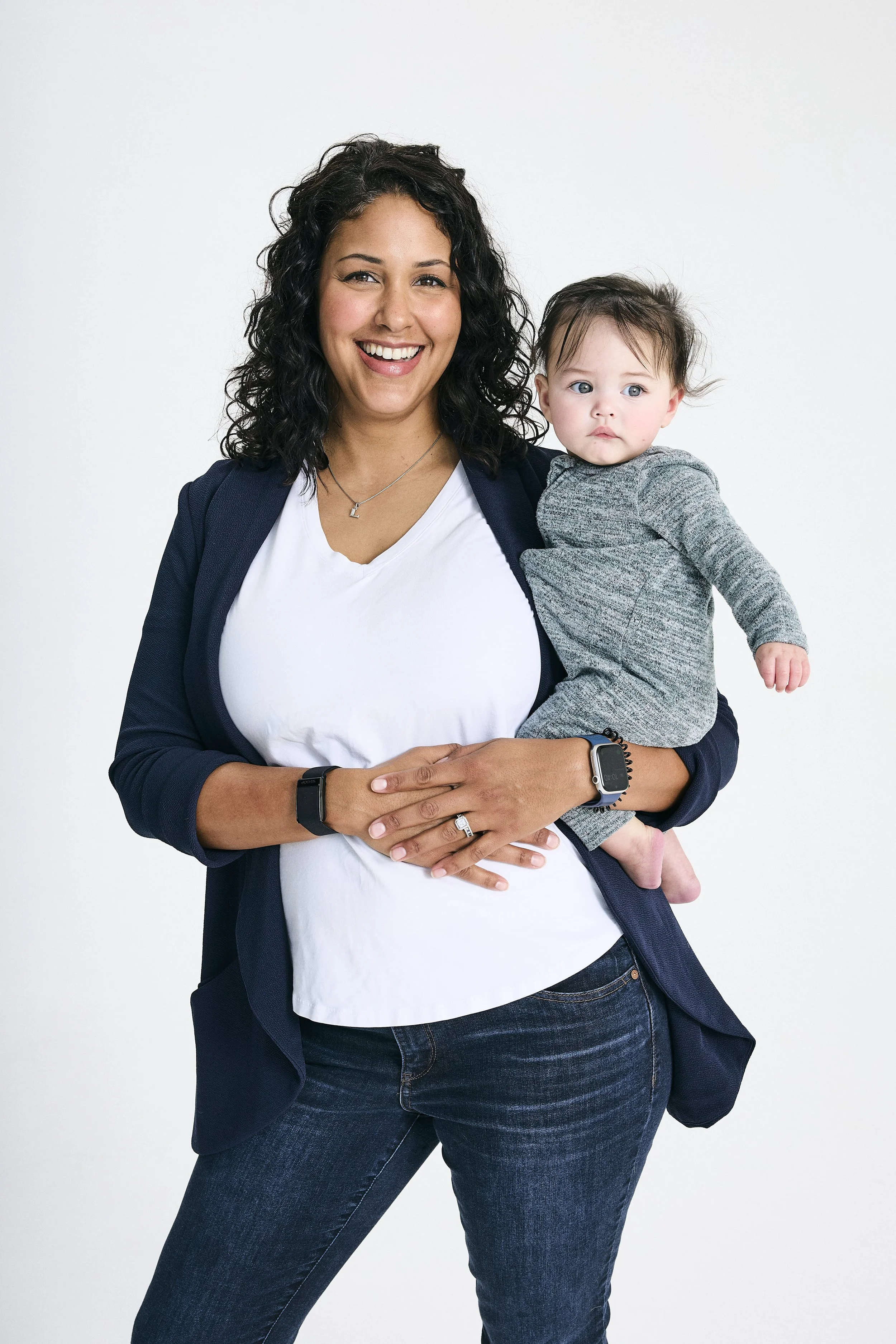 A woman with curly dark hair smiling while holding a small child with blue eyes and brown hair, against a plain white background.