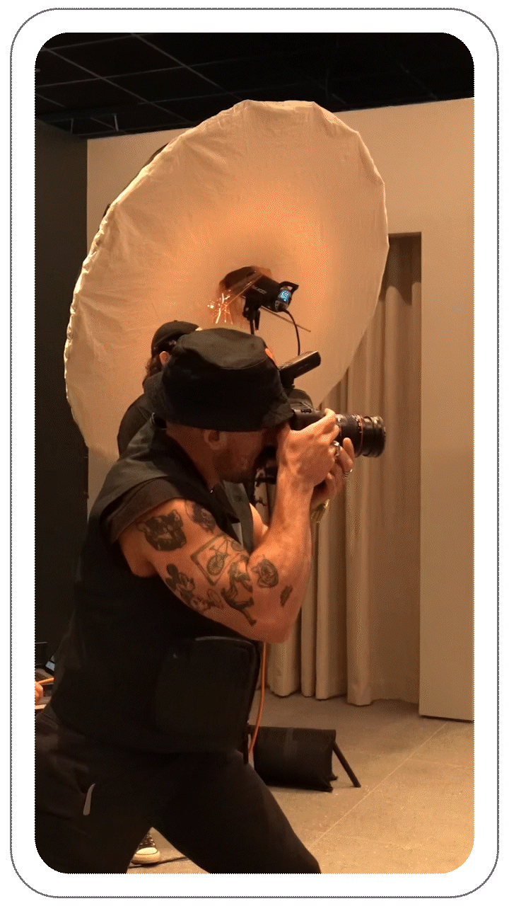 A photographer with tattoos and a bucket hat takes a photo using a camera in a studio, with lighting equipment in the background.