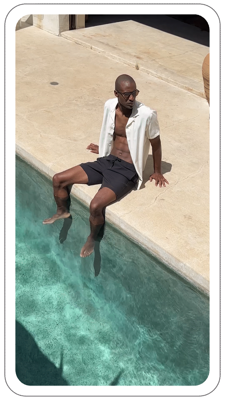 Man sitting on pool edge, feet in water, wearing white shirt and sunglasses.