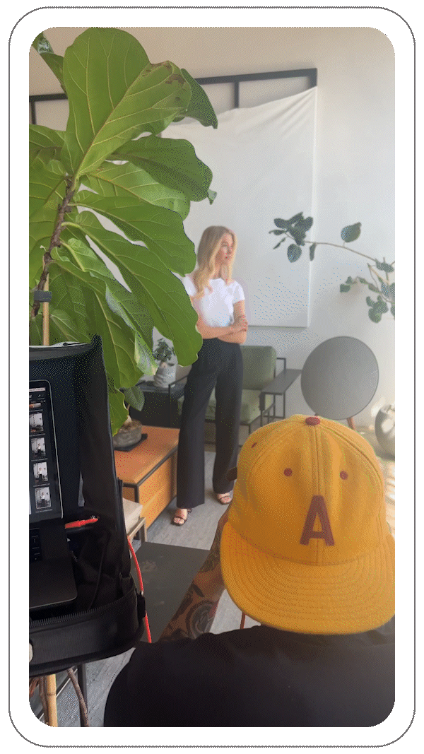 Person in yellow cap photographing a woman posing indoors next to large plants and white backdrop.