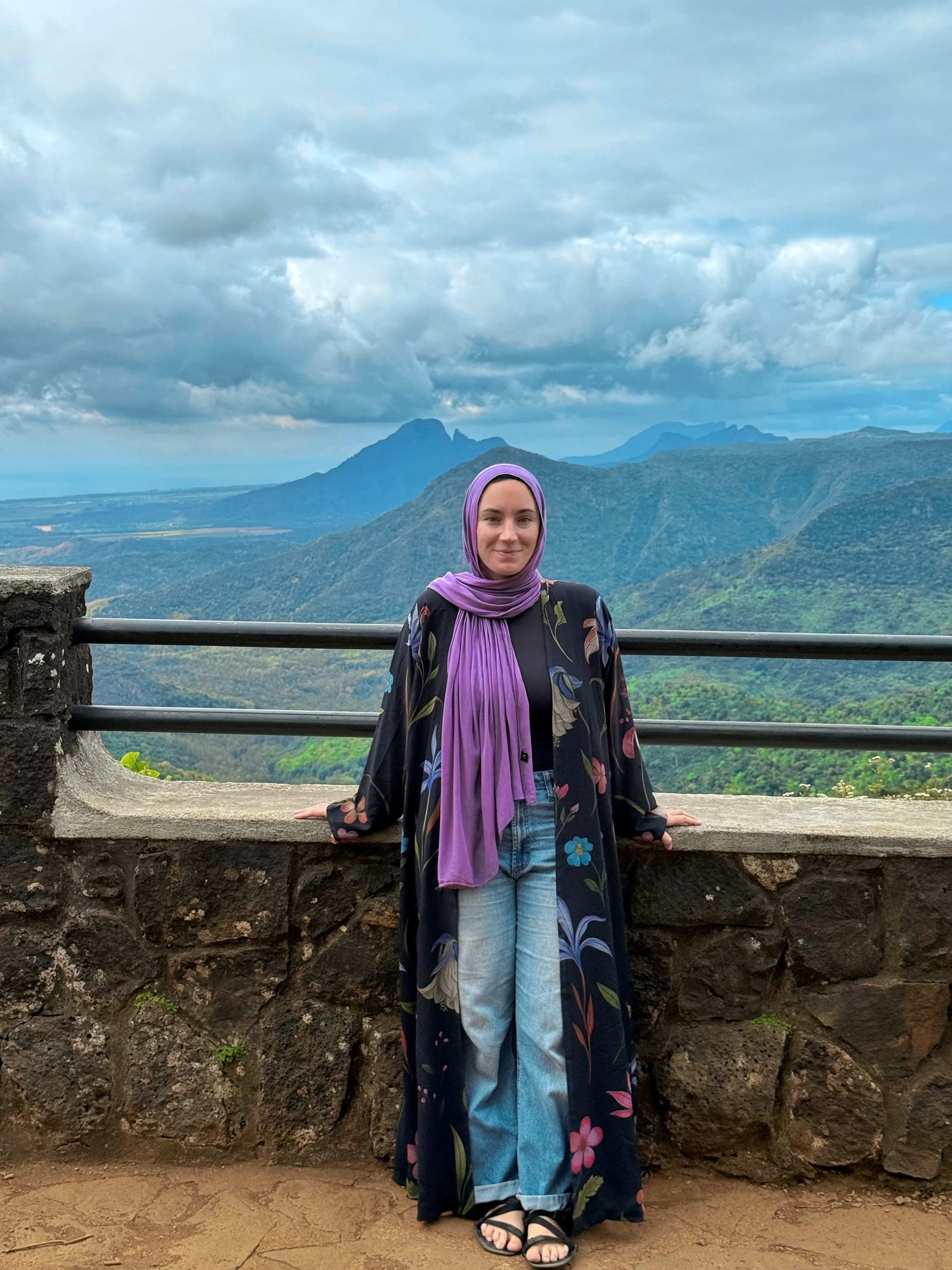 Woman wearing a purple headscarf and a long black floral coat standing by a stone and metal railing, with mountains and cloudy sky in the background.