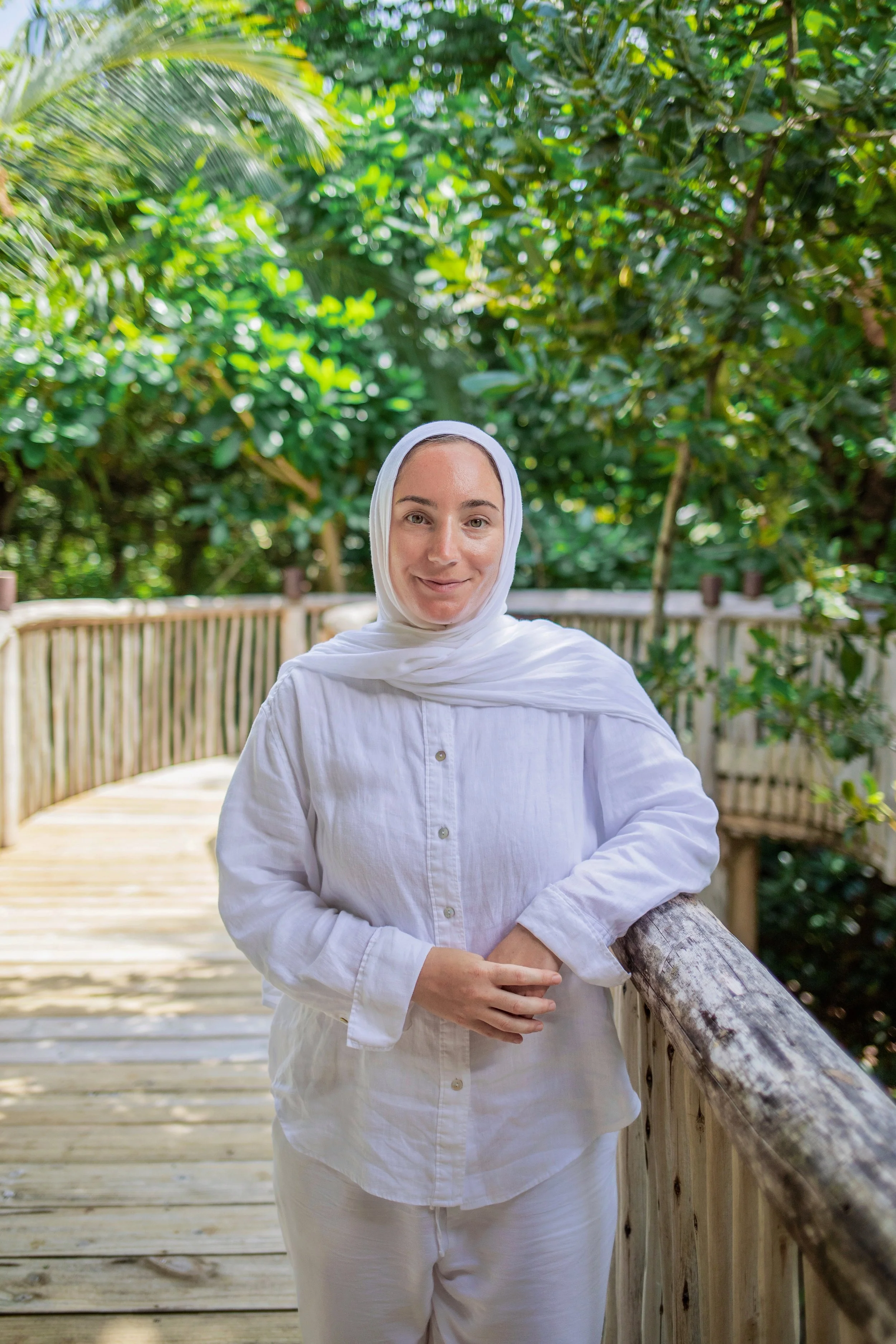 A woman with a white headscarf and white shirt standing on a wooden walkway surrounded by lush green foliage.